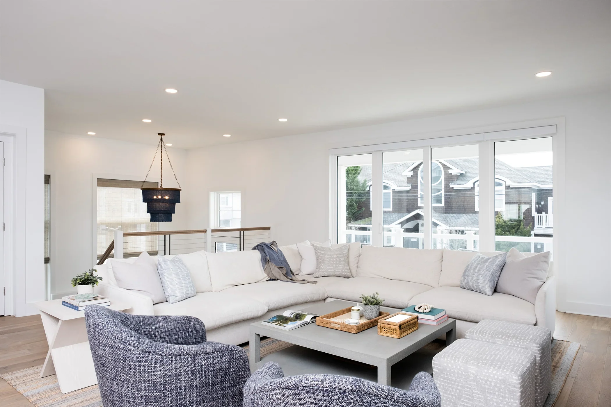 Bright living room with large white sectional sofa, gray armchairs, and a coffee table with books and decor. Behind, large windows reveal neighboring houses. Decorative pillows and a small side table with plants add coziness.