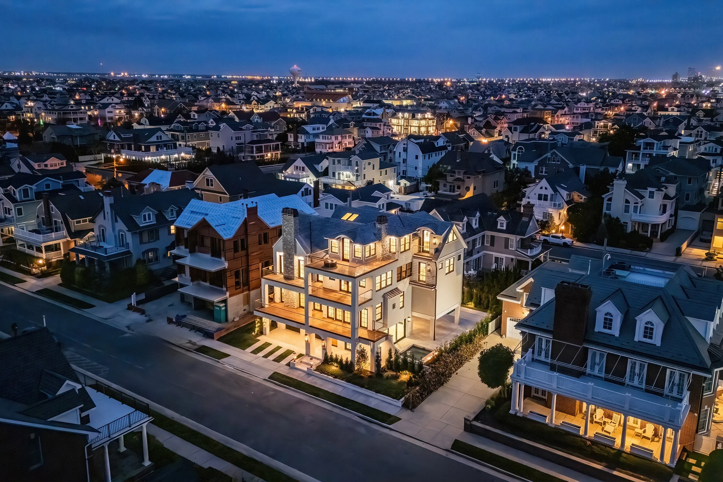 Nighttime aerial view of a residential neighborhood with houses and illuminated windows, showcasing a modern multi-story house with warm lighting on a quiet street.