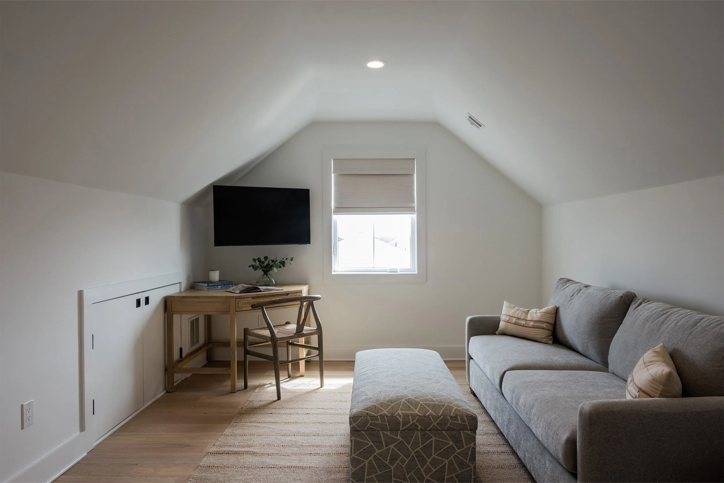 A cozy attic living room with a gray sofa, a patterned ottoman, a small wooden desk with a chair, a window with blinds, and a wall-mounted television.