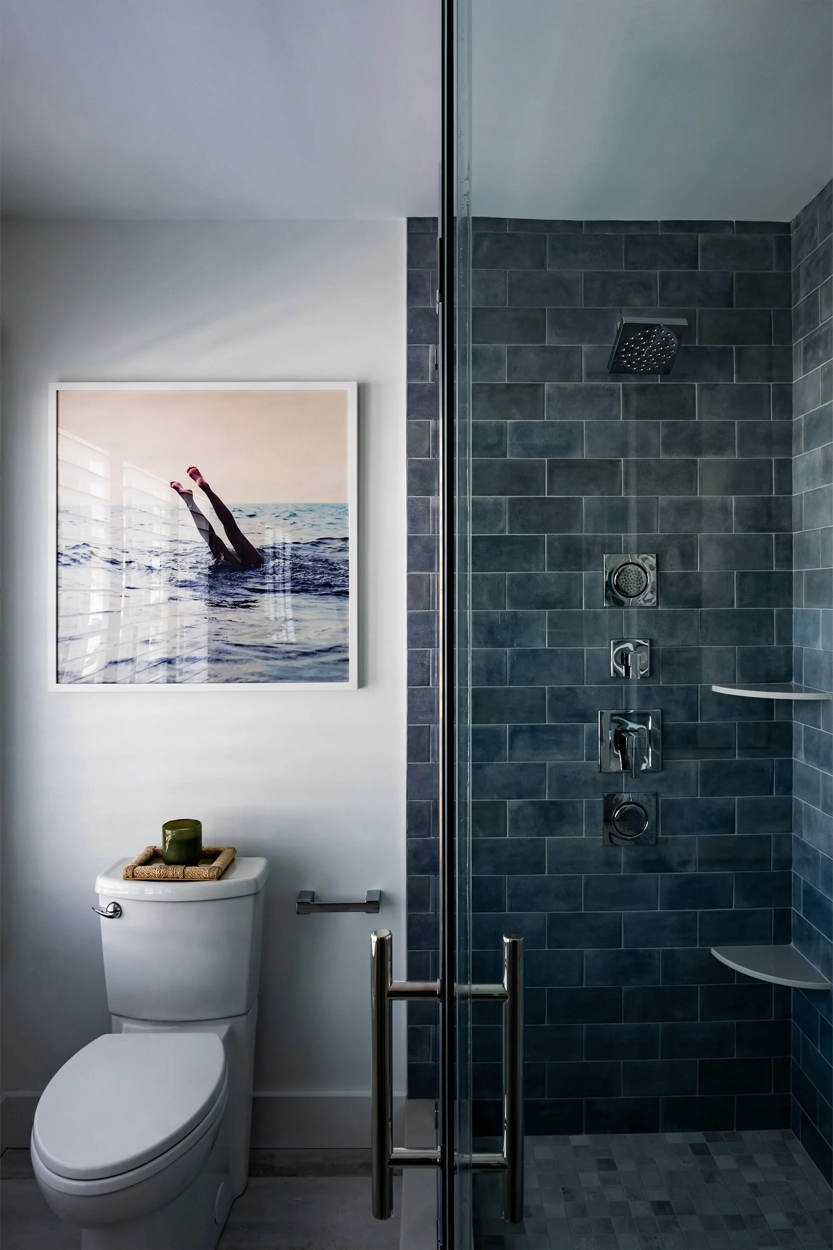 A modern bathroom with a white toilet, decorative items on top, a wall-mounted photo of legs in water, and a glass door to a dark tiled shower area with multiple shower controls and shelves.