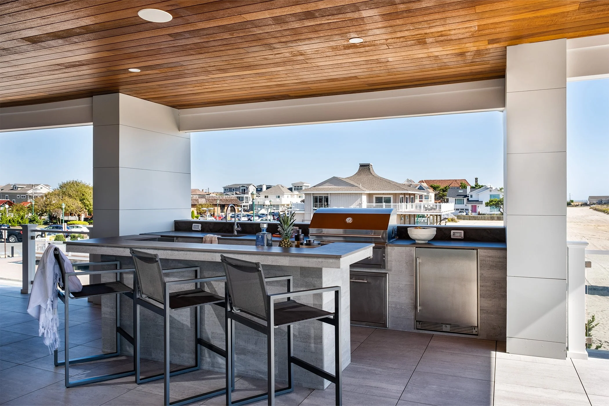 Outdoor kitchen area with a bar counter, two chairs, a grill, a pineapple, bowls, and soap dispenser, with houses and blue sky in the background.