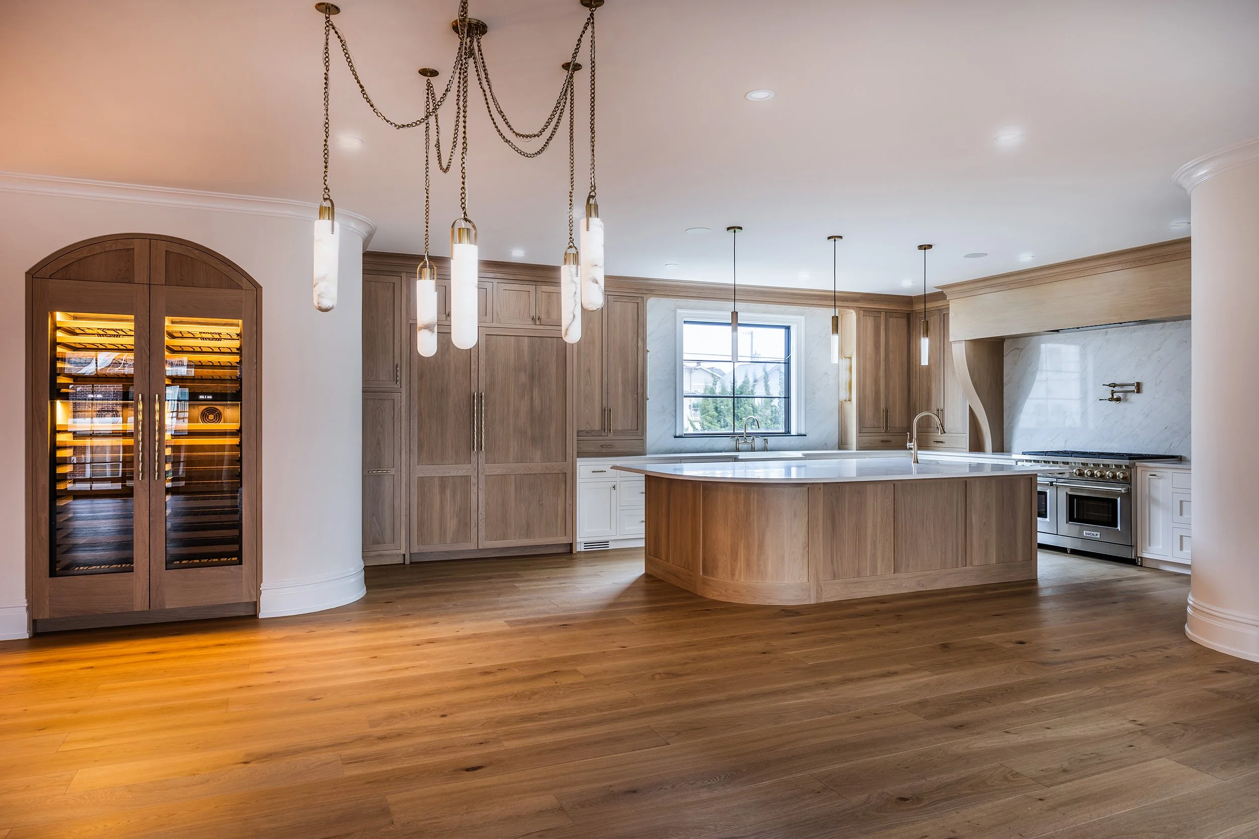 Modern kitchen with wooden cabinets, a kitchen island, a large window, and hardwood floors. There is a wine fridge on the left and a stove with a marble backsplash on the right.