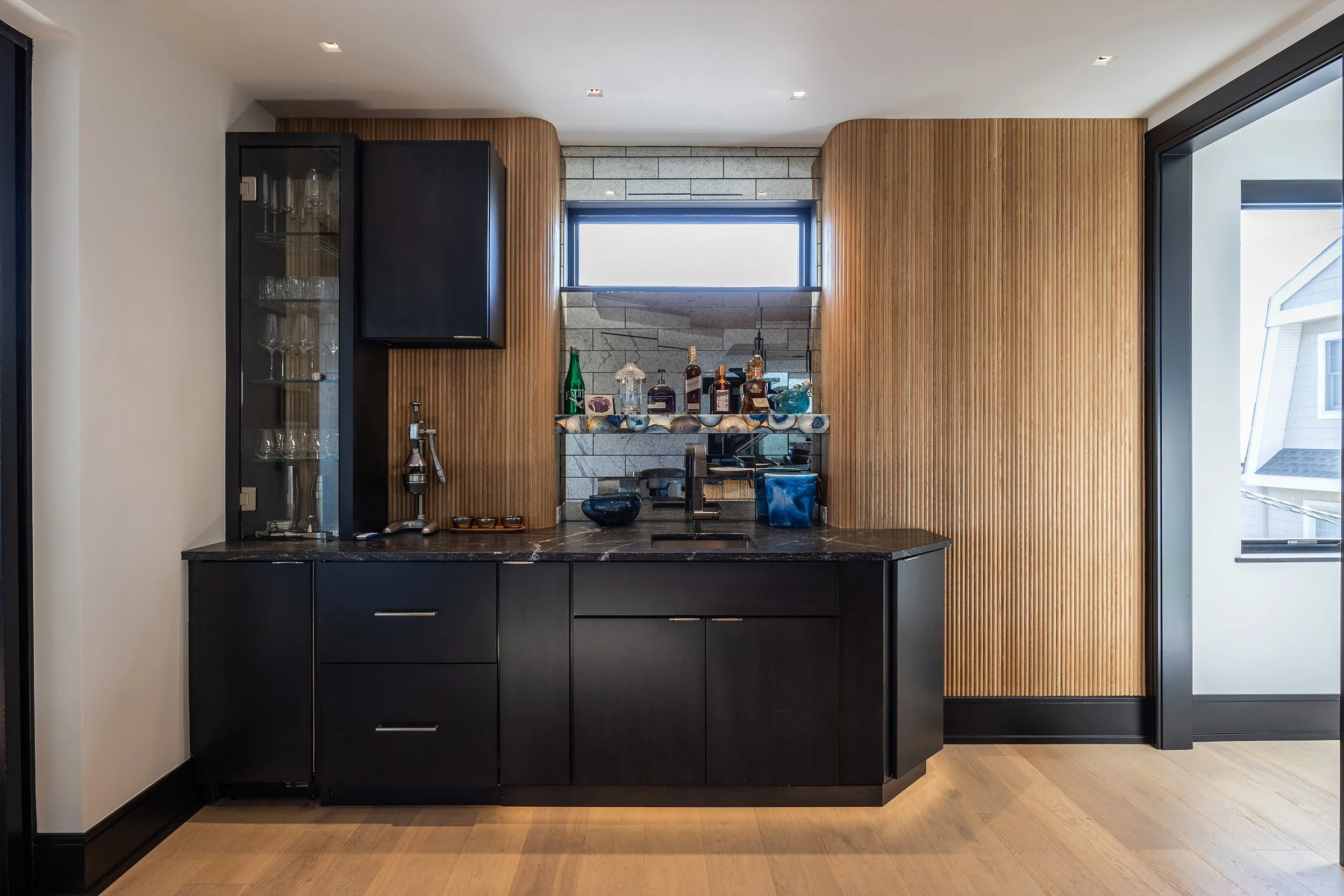 Modern kitchen with black cabinets, a dark countertop, a small sink, wood panel walls, a window with a reflection of the outside, and shelves with bottles and glasses.