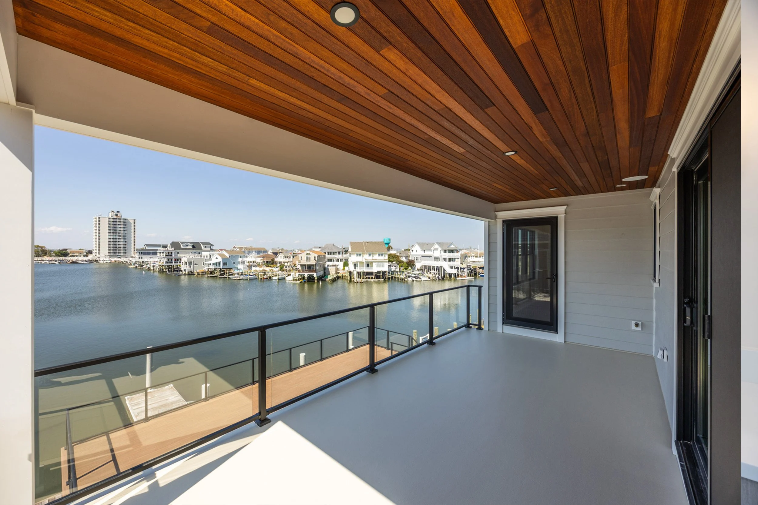 Balcony overlooking waterfront with houses and a tall building in the distance, featuring a wooden ceiling and glass railing.