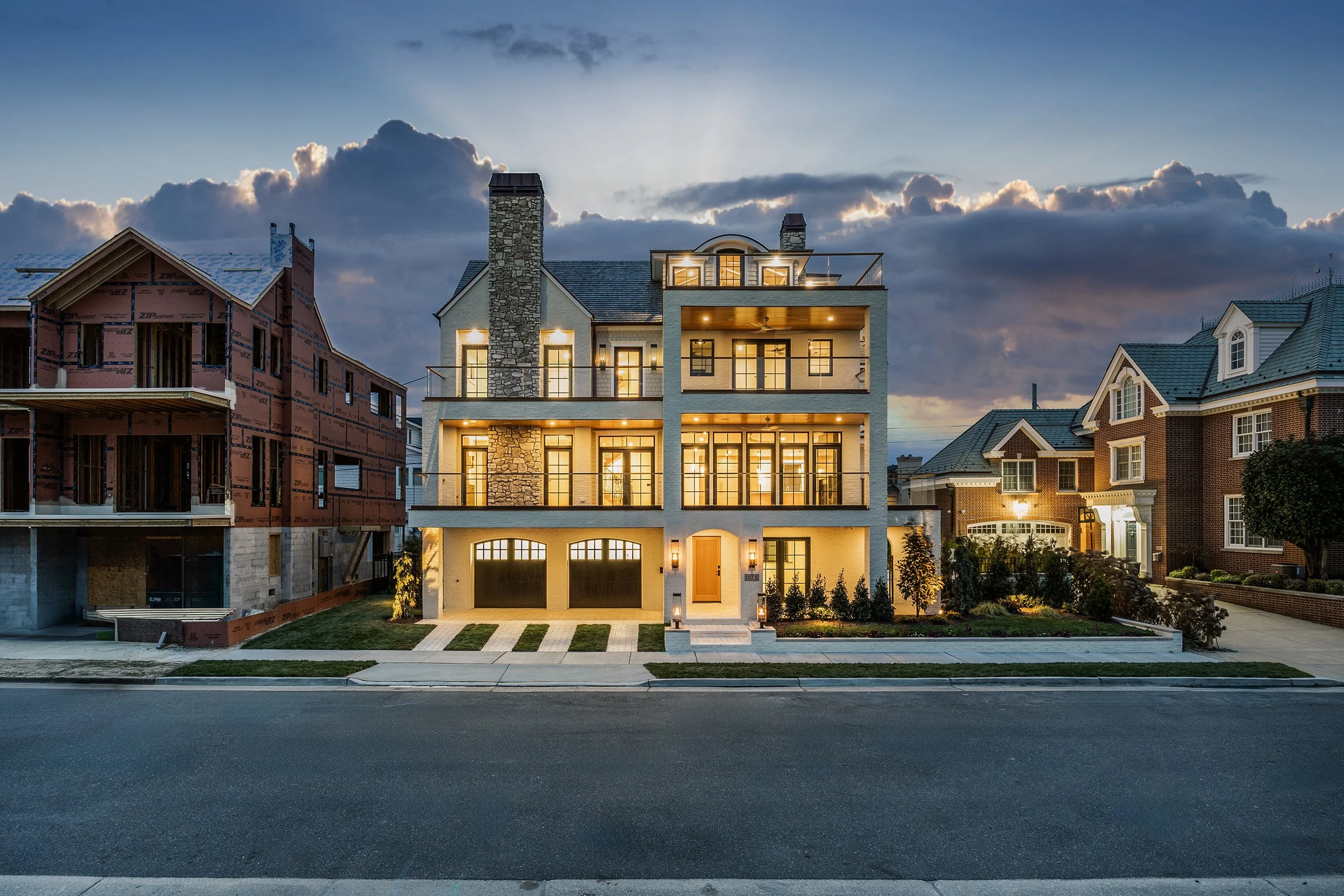 A modern, multi-story house with large glass windows and balconies, illuminated at dusk, situated between a brick house on the right and construction site on the left.