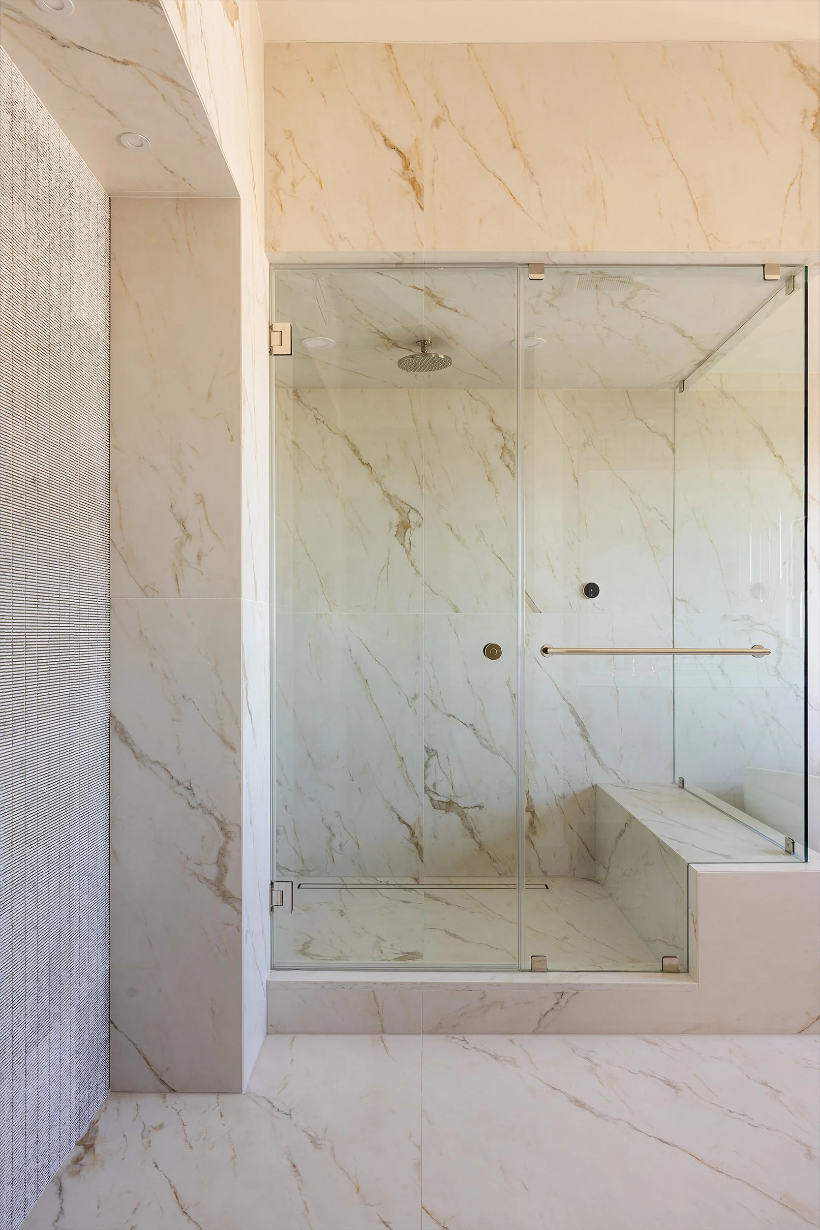 A modern, glass-enclosed shower with marble walls and a built-in bench. The shower features a rainfall showerhead and safety grab bars, with matching marble flooring and a ceiling-mounted light.