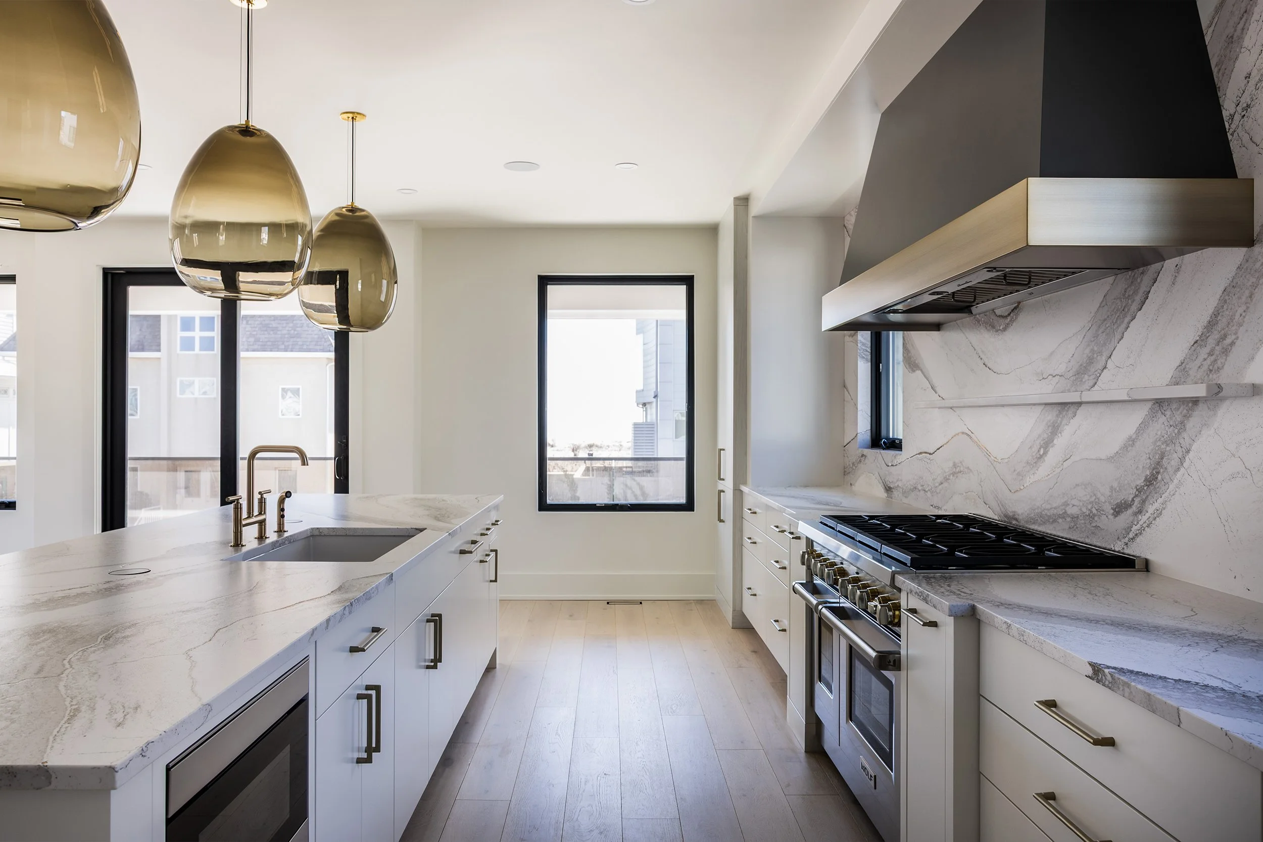 Modern kitchen with white cabinets, marble countertops, and a stainless steel stove. Three gold pendant lights hang above the island. There is a window and a sliding glass door bringing in natural light.