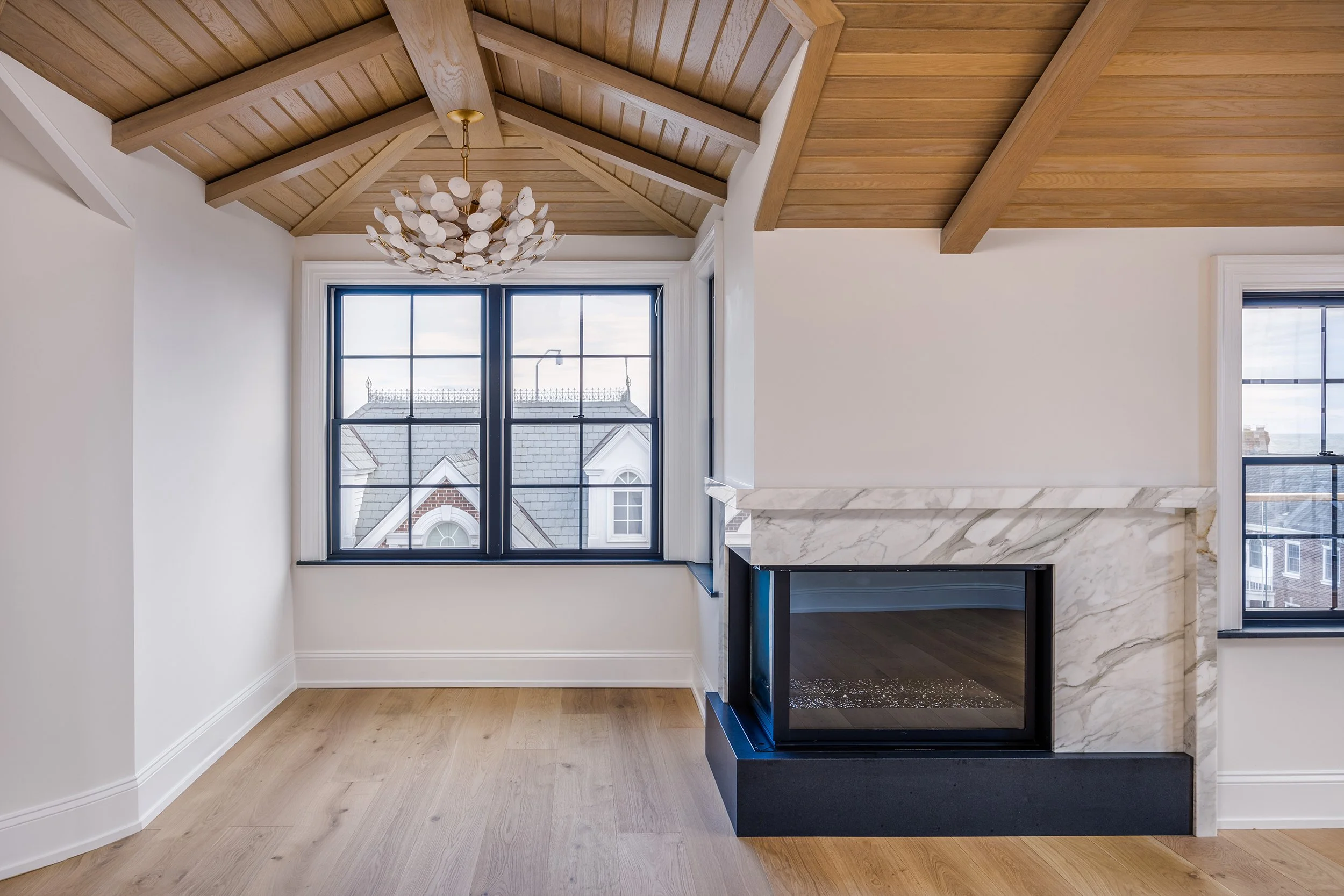 Living room with vaulted wooden ceiling, chandelier, black-framed windows, white walls, and a marble fireplace.