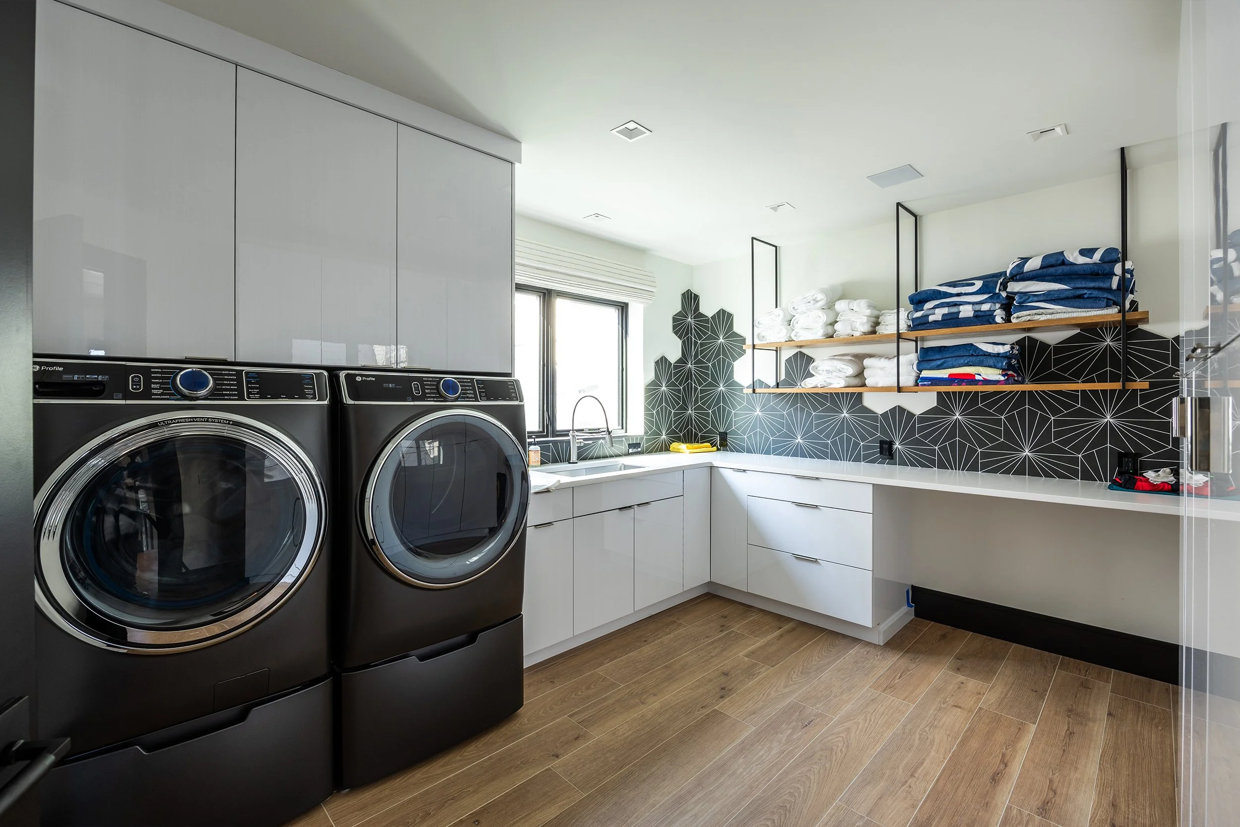 Modern laundry room with front-loading washer and dryer, white cabinets, open shelves with folded towels and jeans, black geometric tile backsplash, wooden flooring, and a window above the sink.