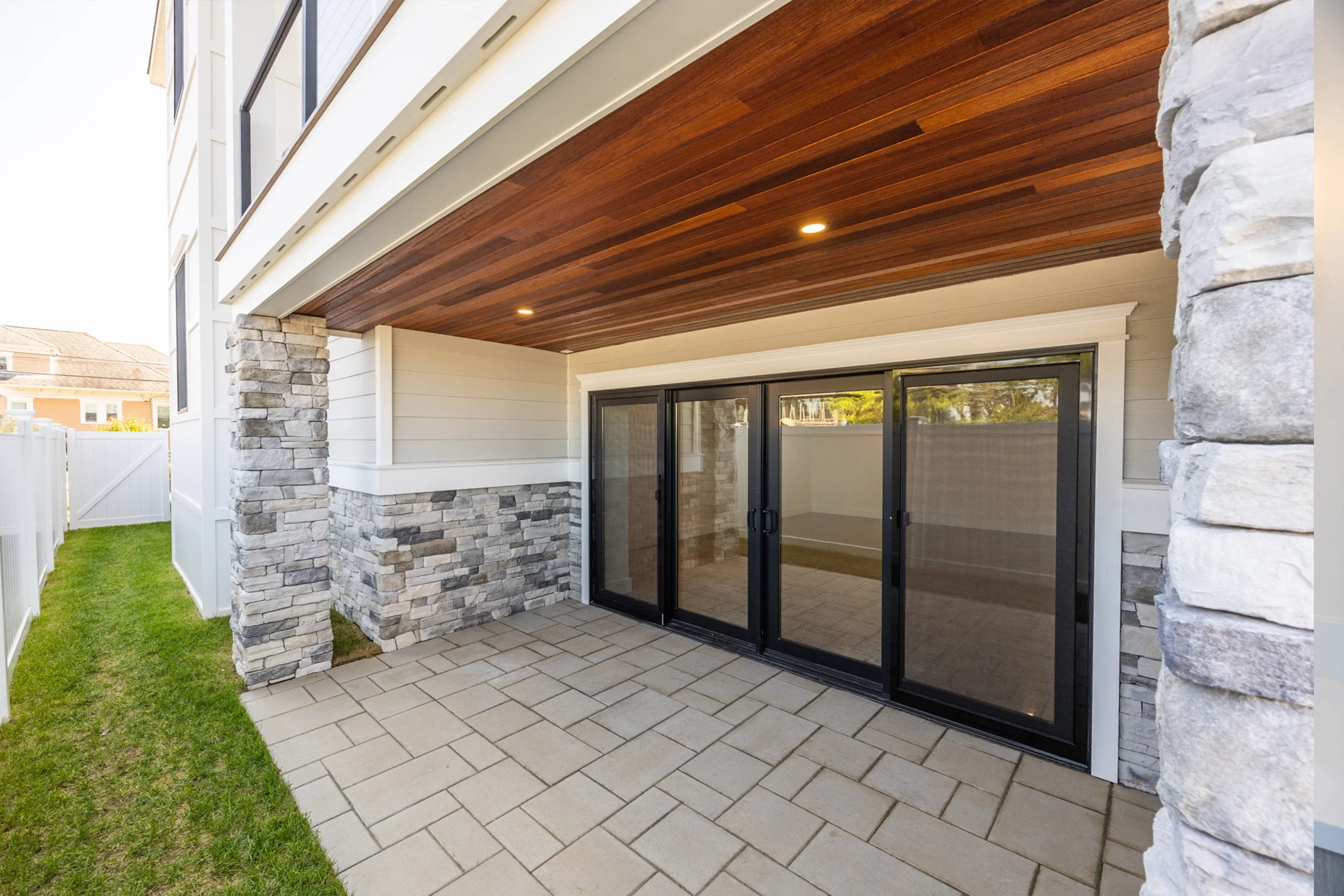 Covered outdoor patio area with beige tile flooring, stone accents on the walls, and sliding glass doors leading inside, with a wooden ceiling and recessed lighting.