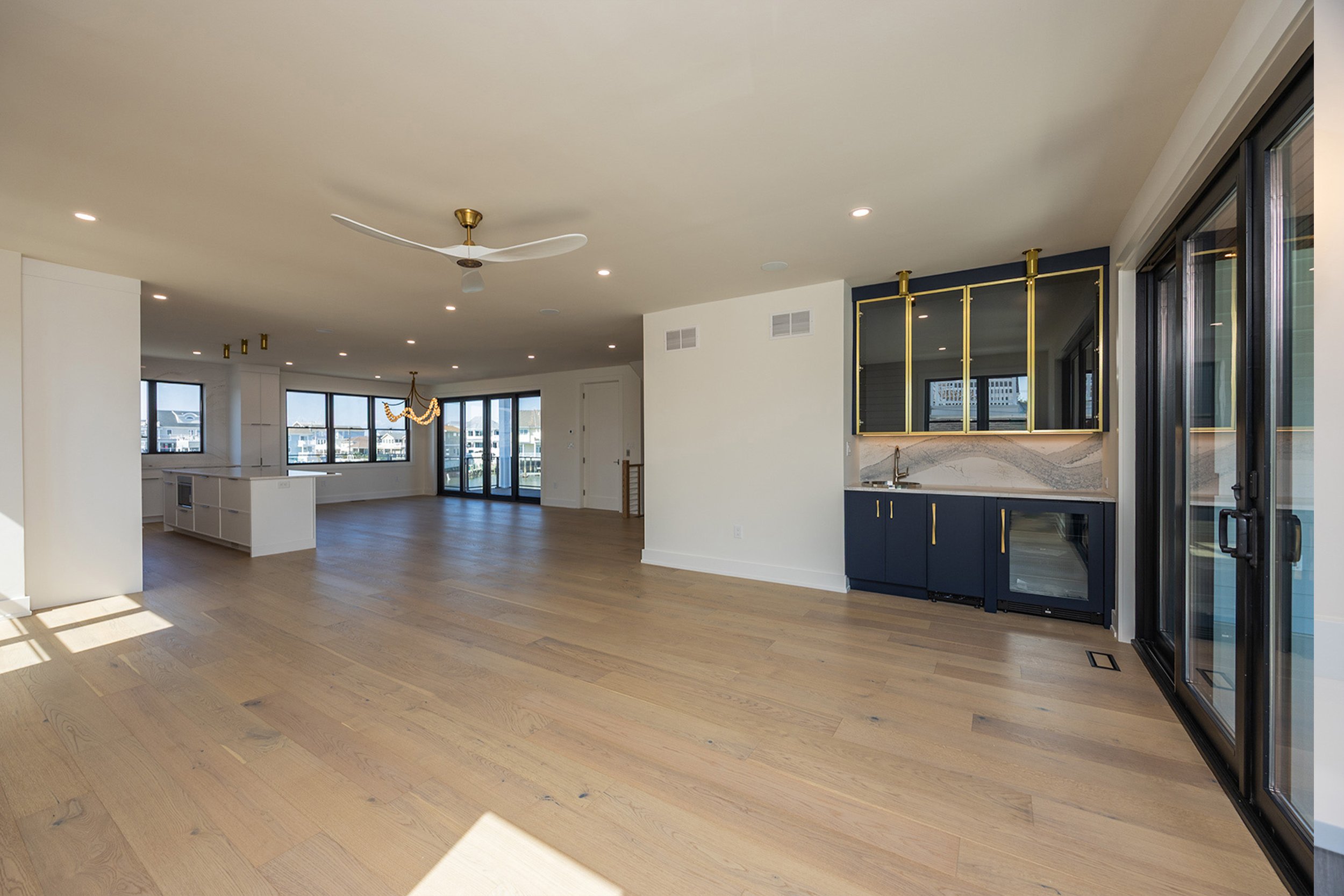 Empty living space with hardwood floors, large windows, and a modern kitchenette with navy blue cabinets and gold accents.