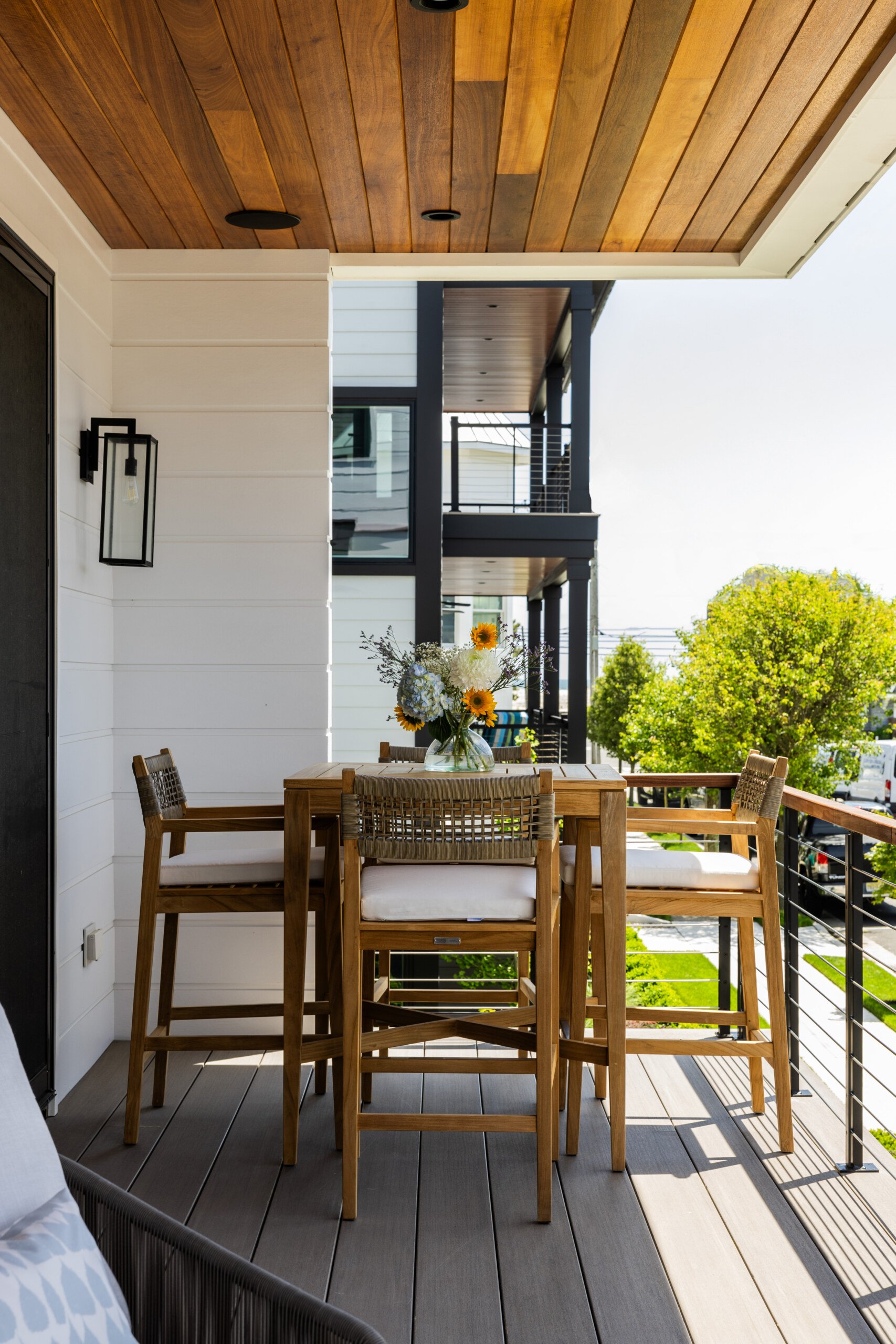 Outdoor balcony with wooden dining table, four chairs with white cushions, a glass vase with a sunflower bouquet, and city trees and buildings in the background.