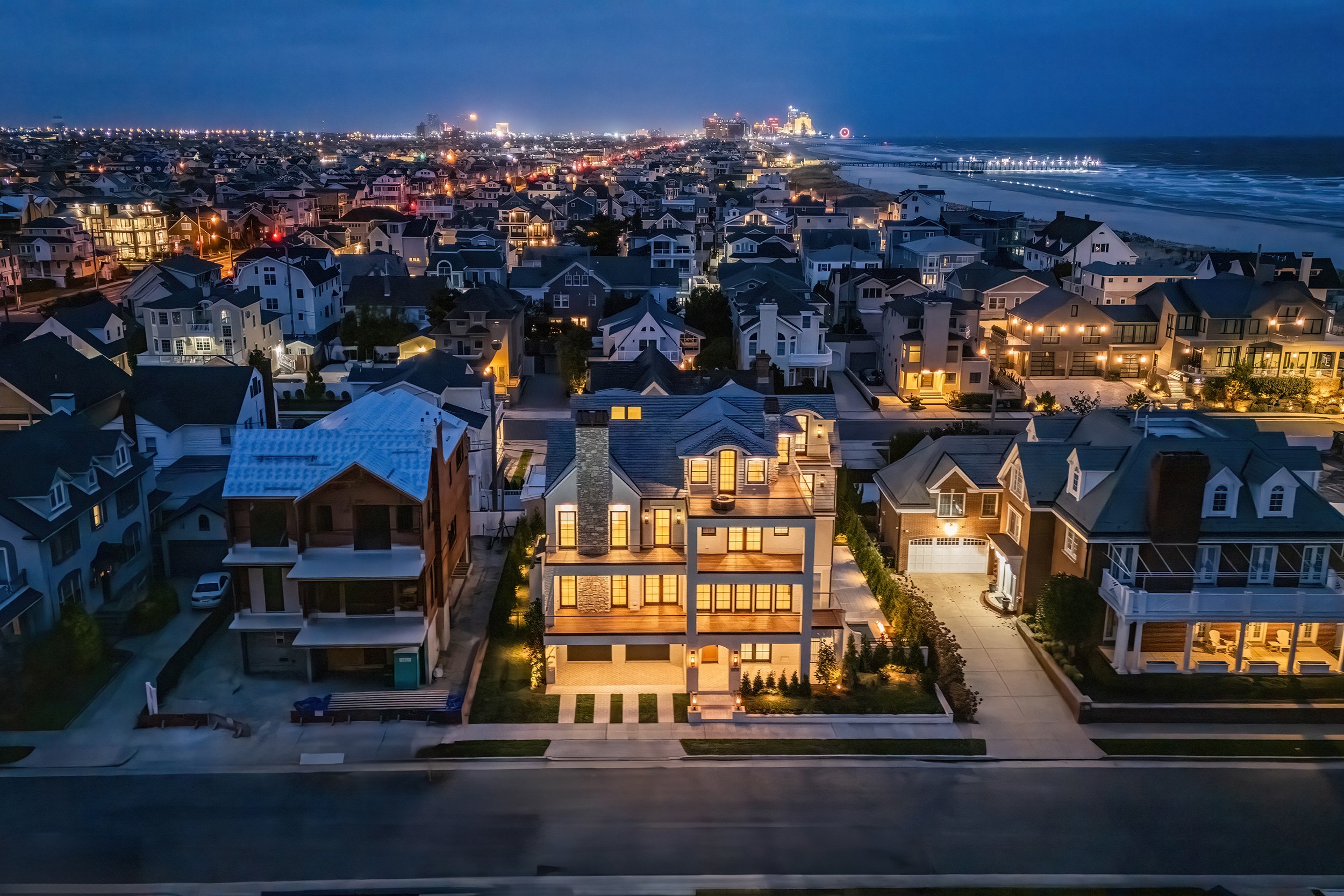 Night view of a coastal residential neighborhood with illuminated modern houses, streets, and an ocean in the background.