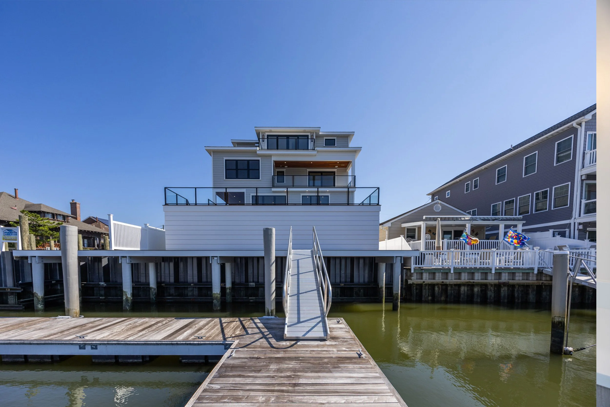 A modern white house with multiple balconies situated on a waterway, with a wooden dock and a metal ramp in the foreground, neighboring houses and clear blue sky in the background.