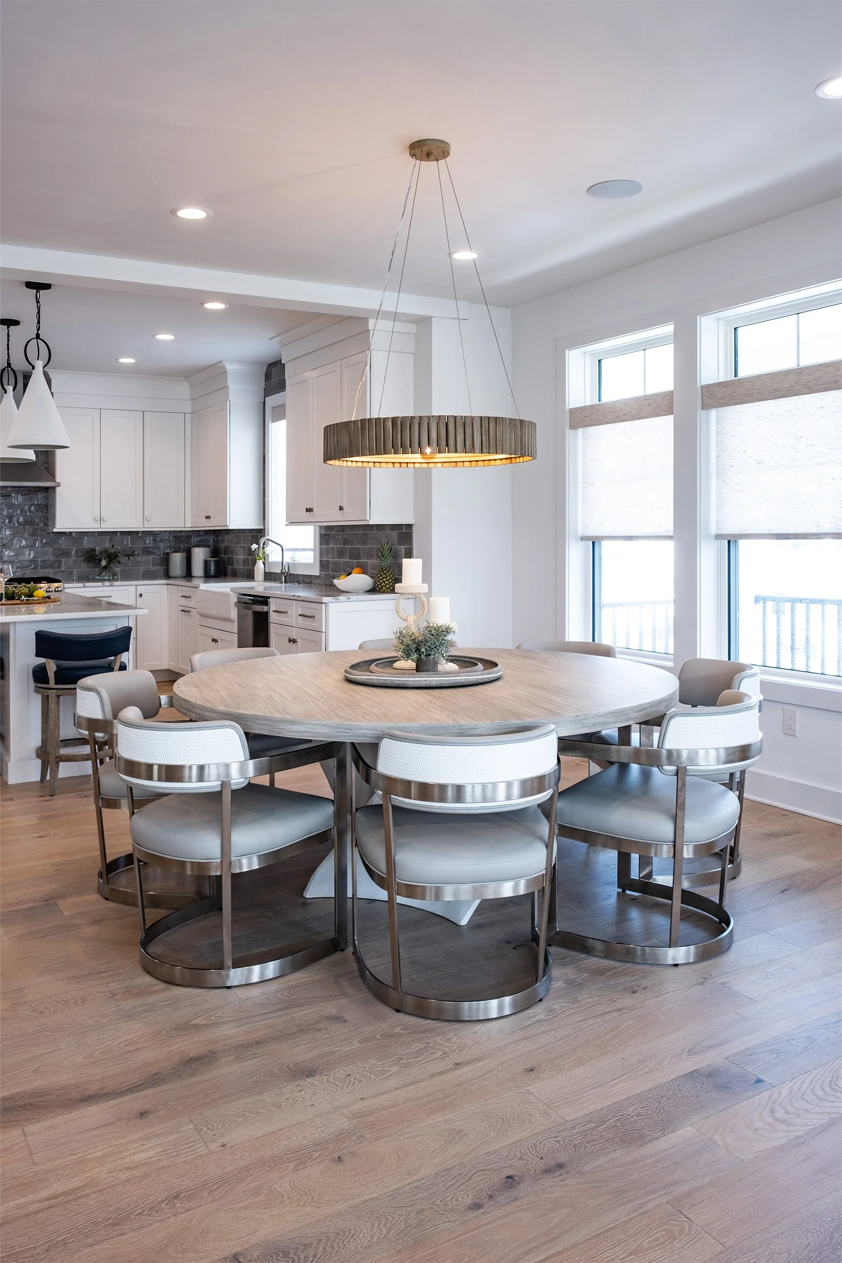 Modern kitchen and dining area with a round wooden table, eight chairs with metal frames and white cushions, a pendant light, and large windows with blinds.