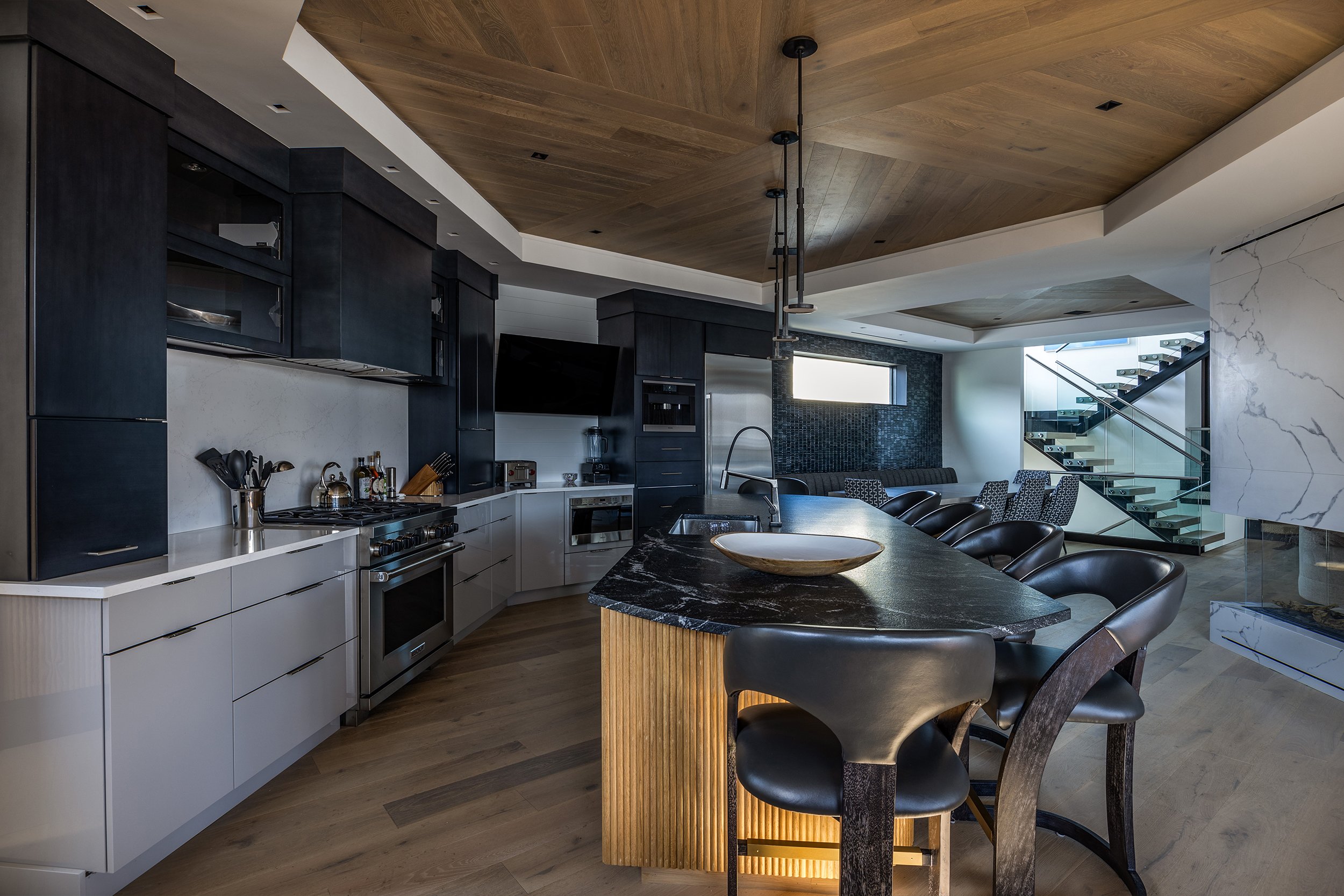 Modern kitchen with black and white cabinets, a black marble island with a wooden base, black chairs, and a view of a living area with staircase and large window.