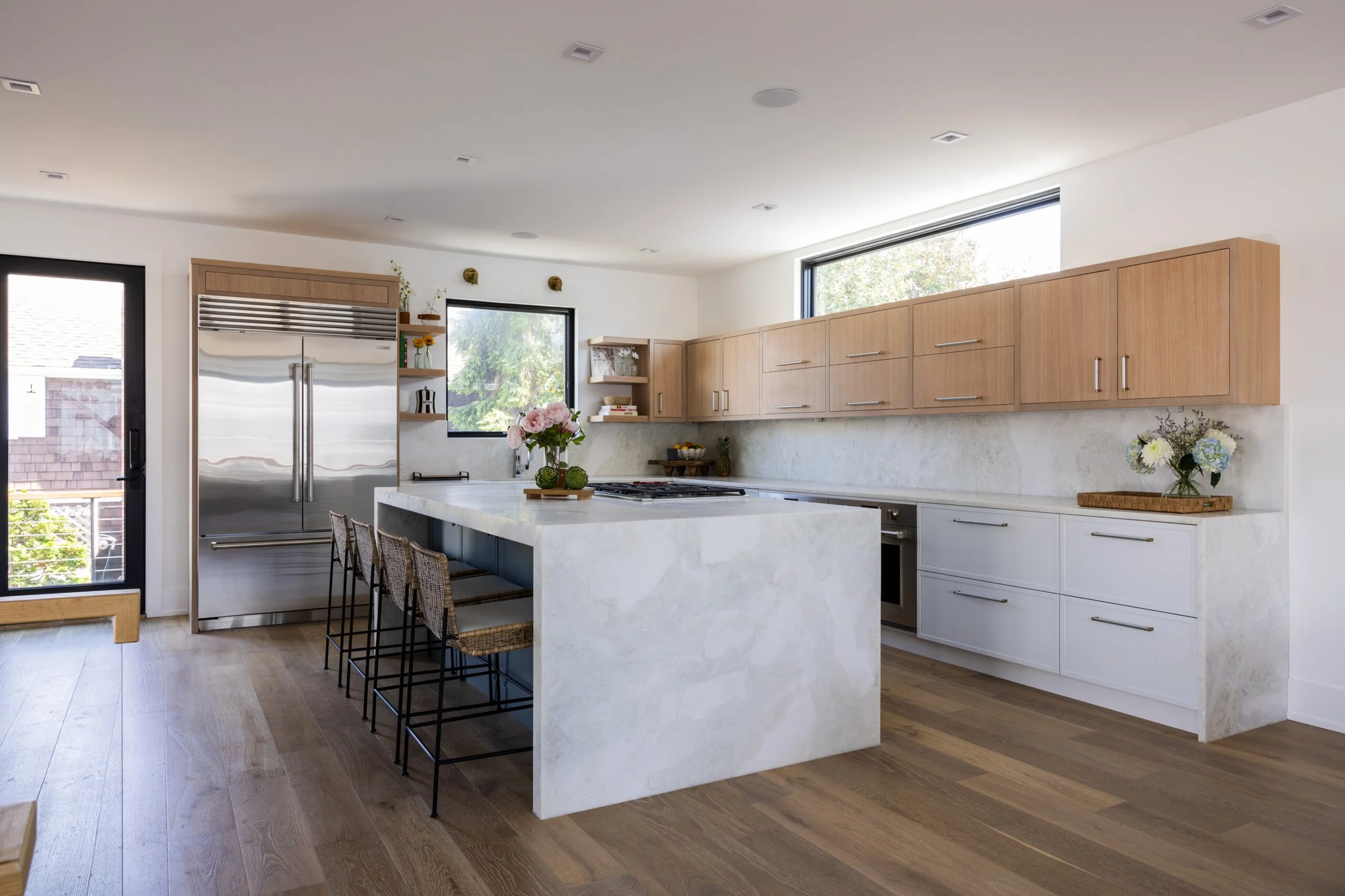 Modern kitchen with white and wood cabinetry, a large marble island with bar stools, a stainless steel refrigerator, and natural light from windows and a glass door.