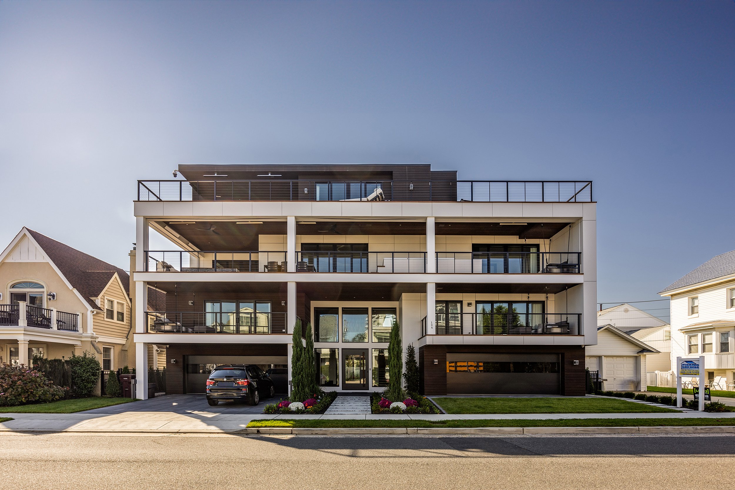 Multiple-story modern apartment building with balconies, large glass doors, and a landscaped front yard with bushes and flowers, situated in a suburban neighborhood.