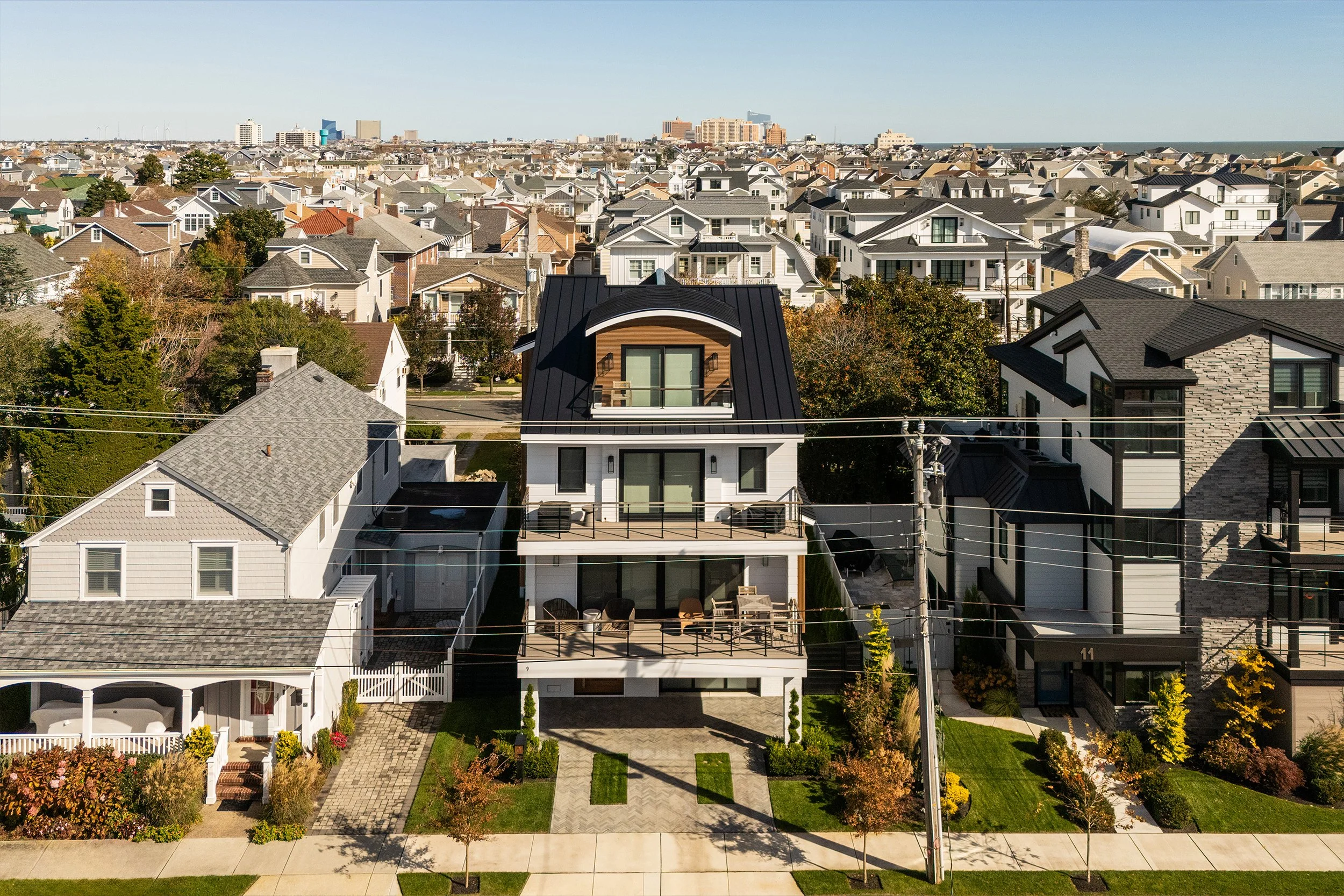 Aerial view of a modern residential neighborhood with a large, white, multi-story house featuring a black roof, surrounded by smaller houses, trees, and a sidewalk.