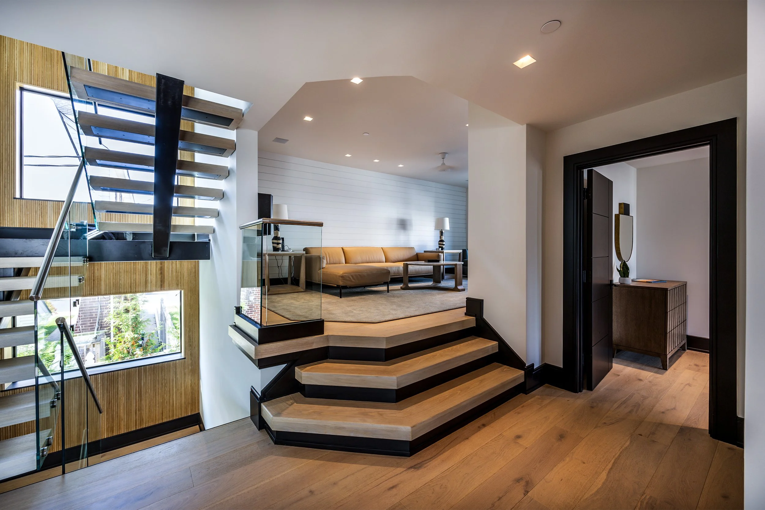 Interior view of a modern home staircase leading to a living room with a tan sofa, side tables, lamps, and a rug. The space is well-lit with recessed lighting and has a window on the staircase wall.