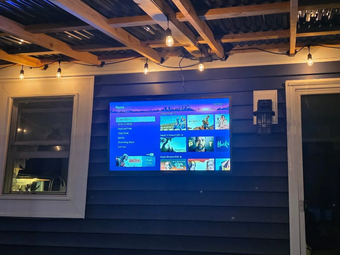A flat-screen TV mounted on the exterior of a house, displaying a streaming service menu, with string lights hanging from a wooden ceiling above, near a window and a door.