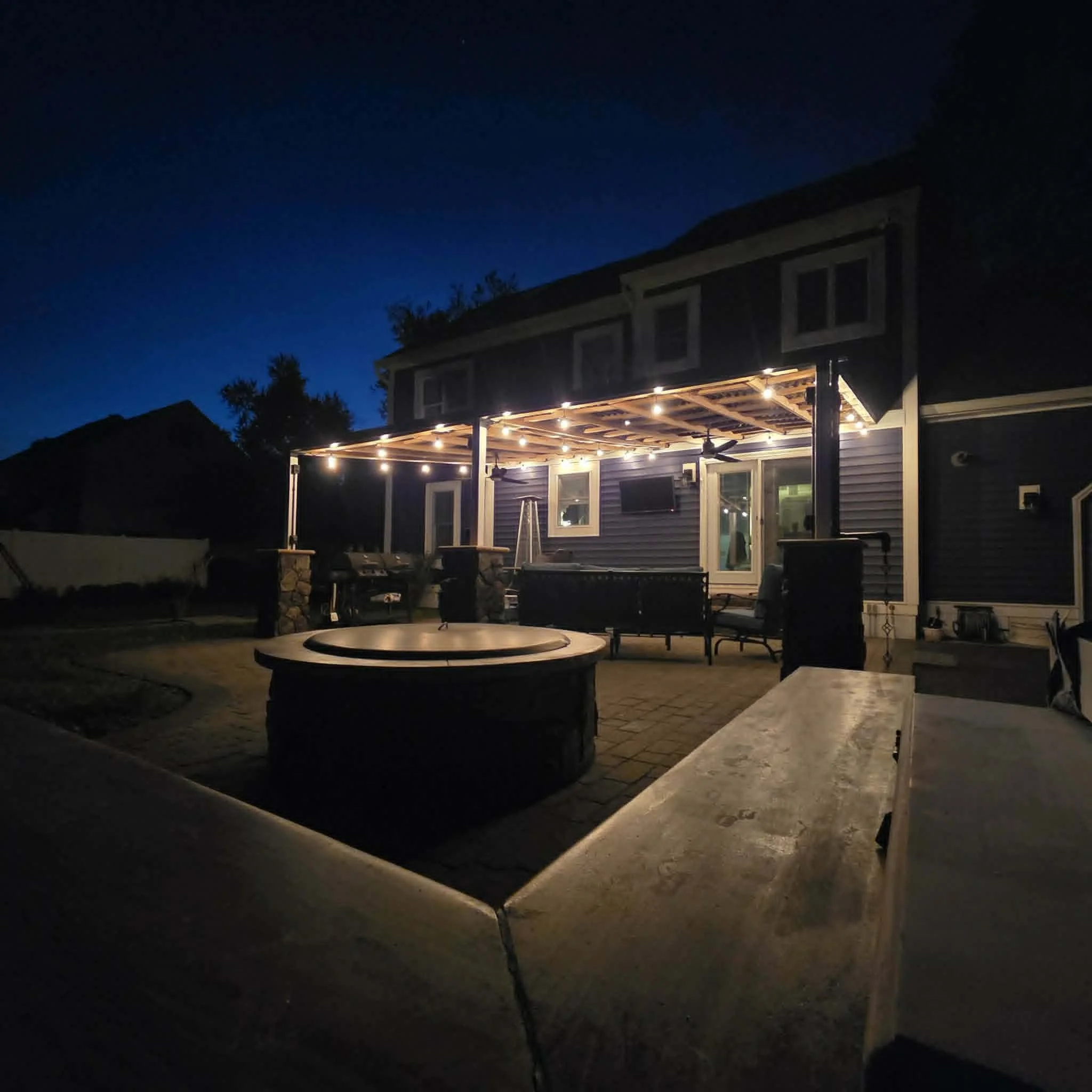 A house with outdoor string lights hanging from the porch. The house has blue siding and several windows, with a dark evening sky overhead.