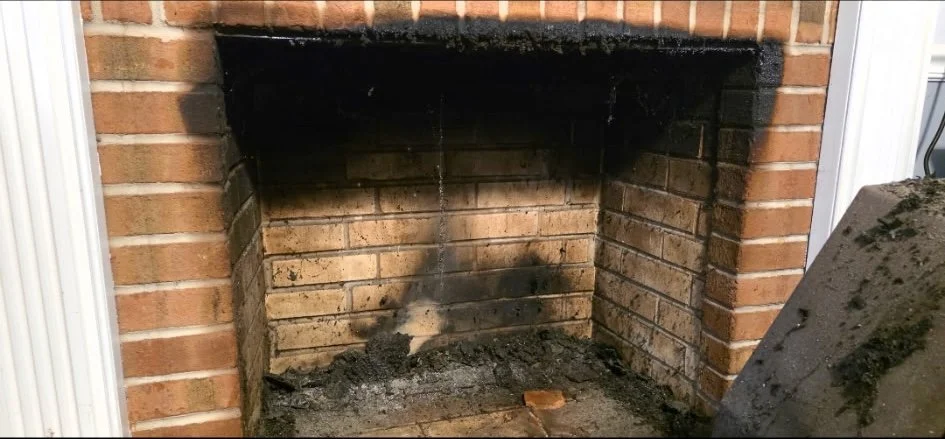 The image shows a burnt and blackened fireplace with soot stains on the brick interior, a small pile of ashes and debris at the bottom, and a brick wall surround. There is a piece of wood or brick inside the fireplace and part of a nearby wall or app