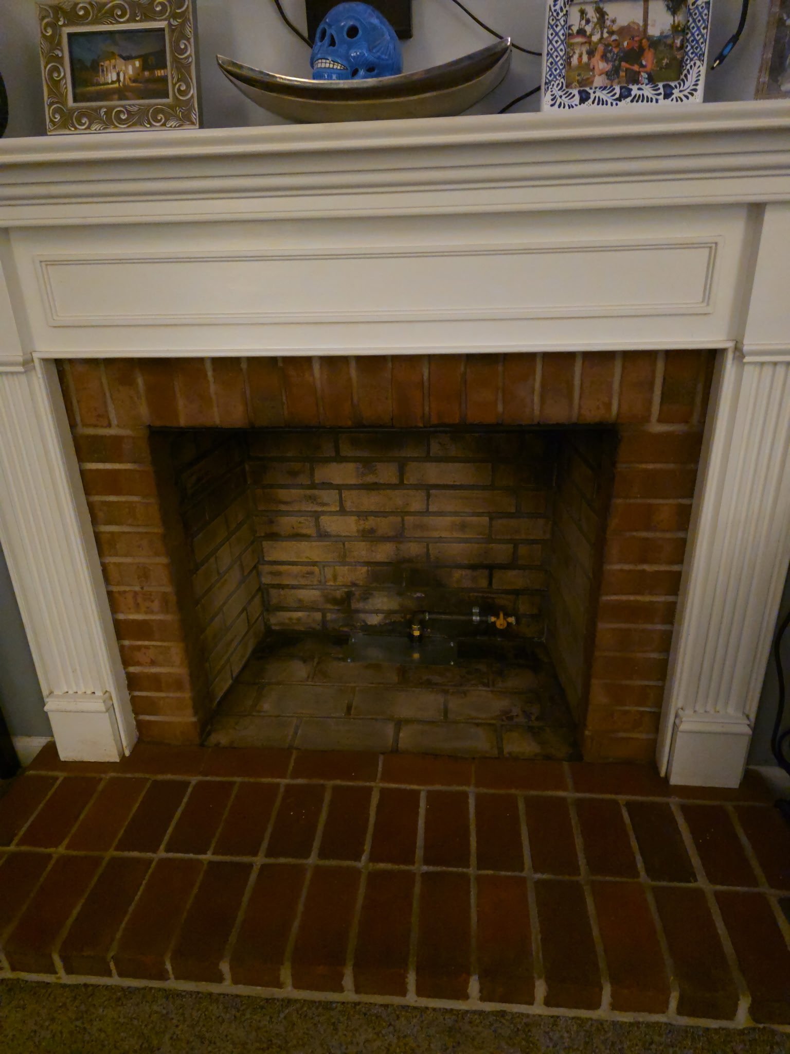 Empty brick fireplace with white mantel, decorative items on top, and framed photos in the background.