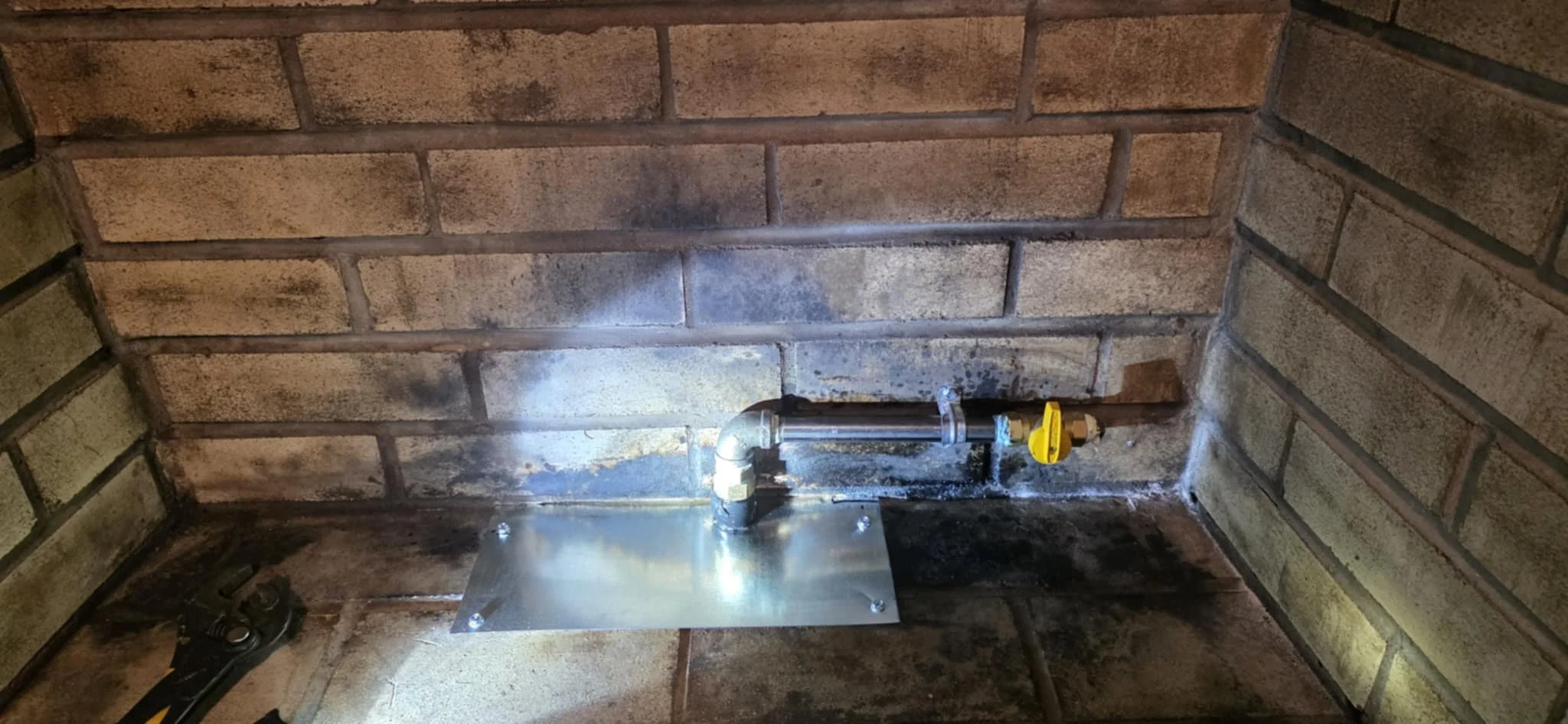 A close-up view of plumbing pipes and a metal plate against a brick wall, with a wrench visible on the floor.