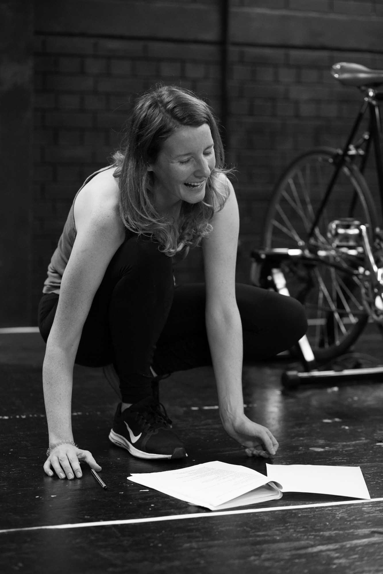 A woman with long hair in athletic clothing and running shoes appears to be laughing or smiling while crouched on the floor, reading or looking at a document or script, with a pen in her hand, near a bicycle.