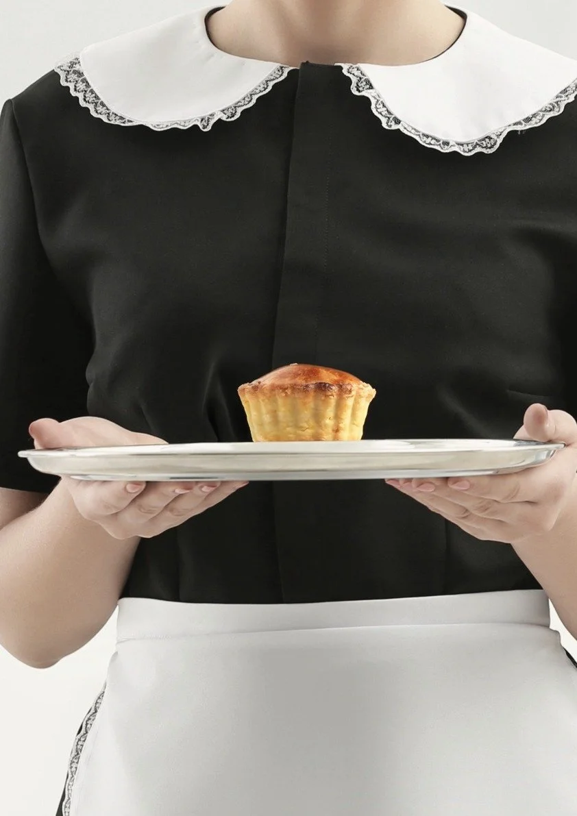 A woman dressed as a waitress or maid in a black and white uniform with a lace-collared top holding a white plate with a baked good, possibly a muffin or tart, in her hands.