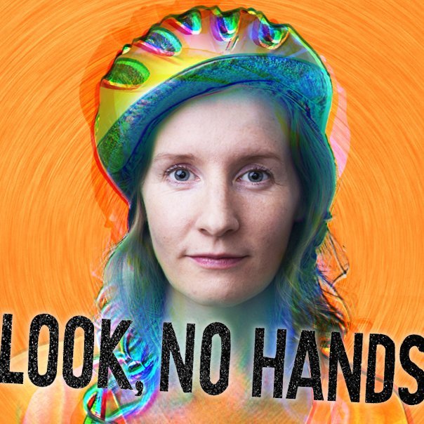 A woman sitting on a stationary bicycle in front of a wall decorated with hanging bicycle wheels and shadows, in a theater or performance space.