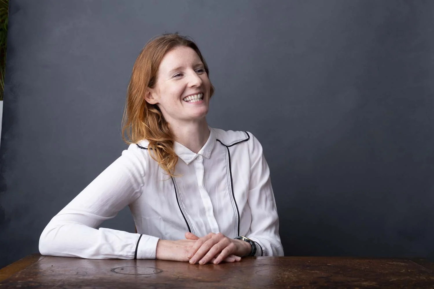 A woman with red hair smiling and sitting at a wooden table against a dark gray background.