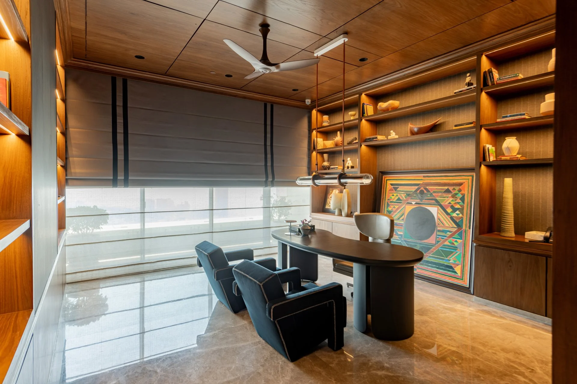 A modern office with wooden bookshelves on the wall, a large window with blinds, a ceiling fan, and a desk with chairs.