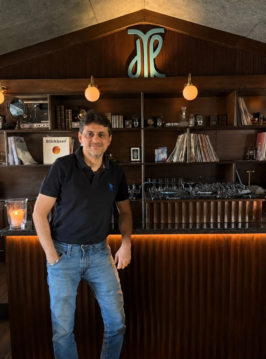 A man in a black polo shirt and jeans standing inside a bar or music lounge with shelves of records, vintage cameras, and records on display behind him, along with a DJ setup.