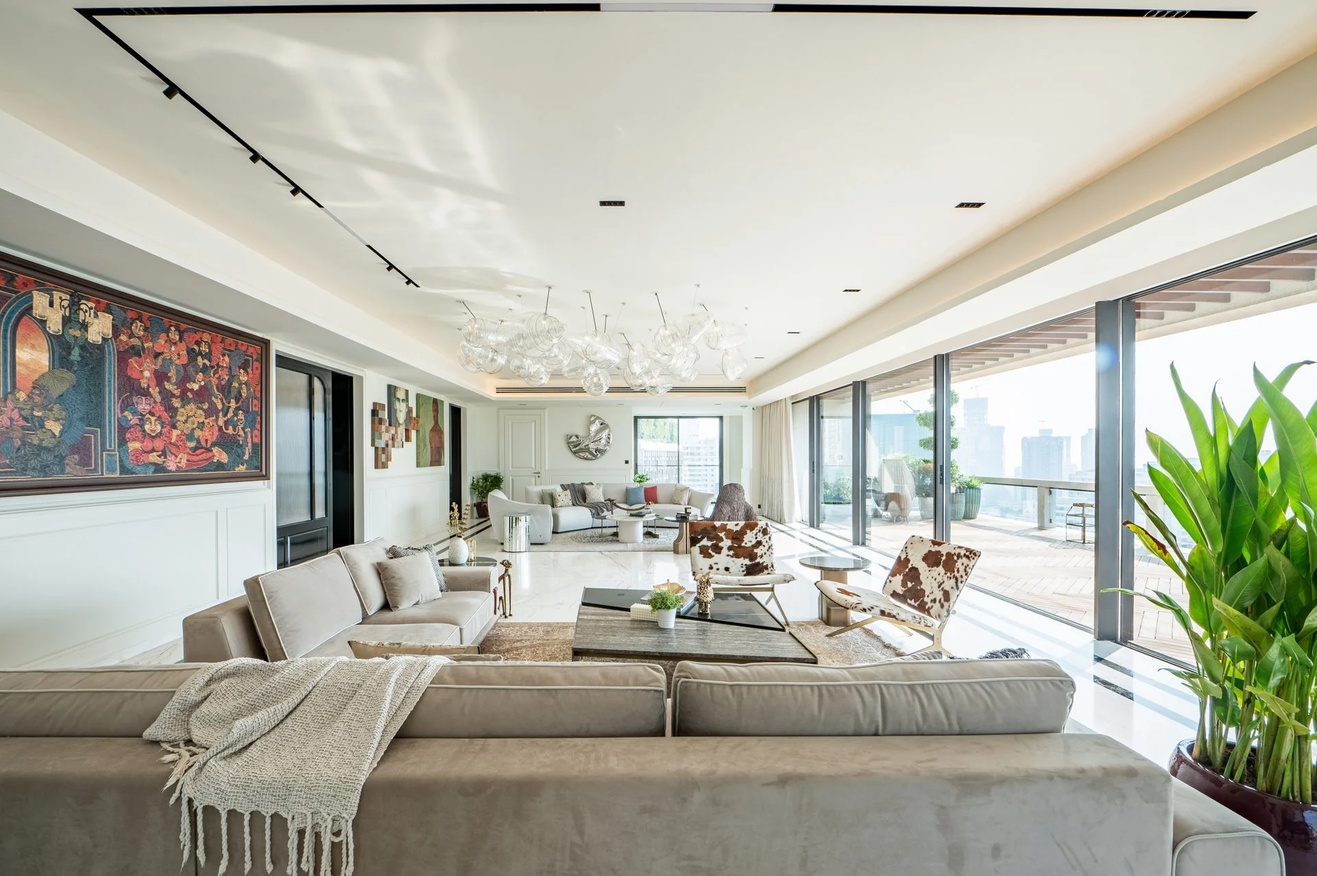 Modern spacious living room with beige sofa, cowhide chairs, abstract artwork, glass chandelier, large windows, balcony with outdoor furniture, and green plants.