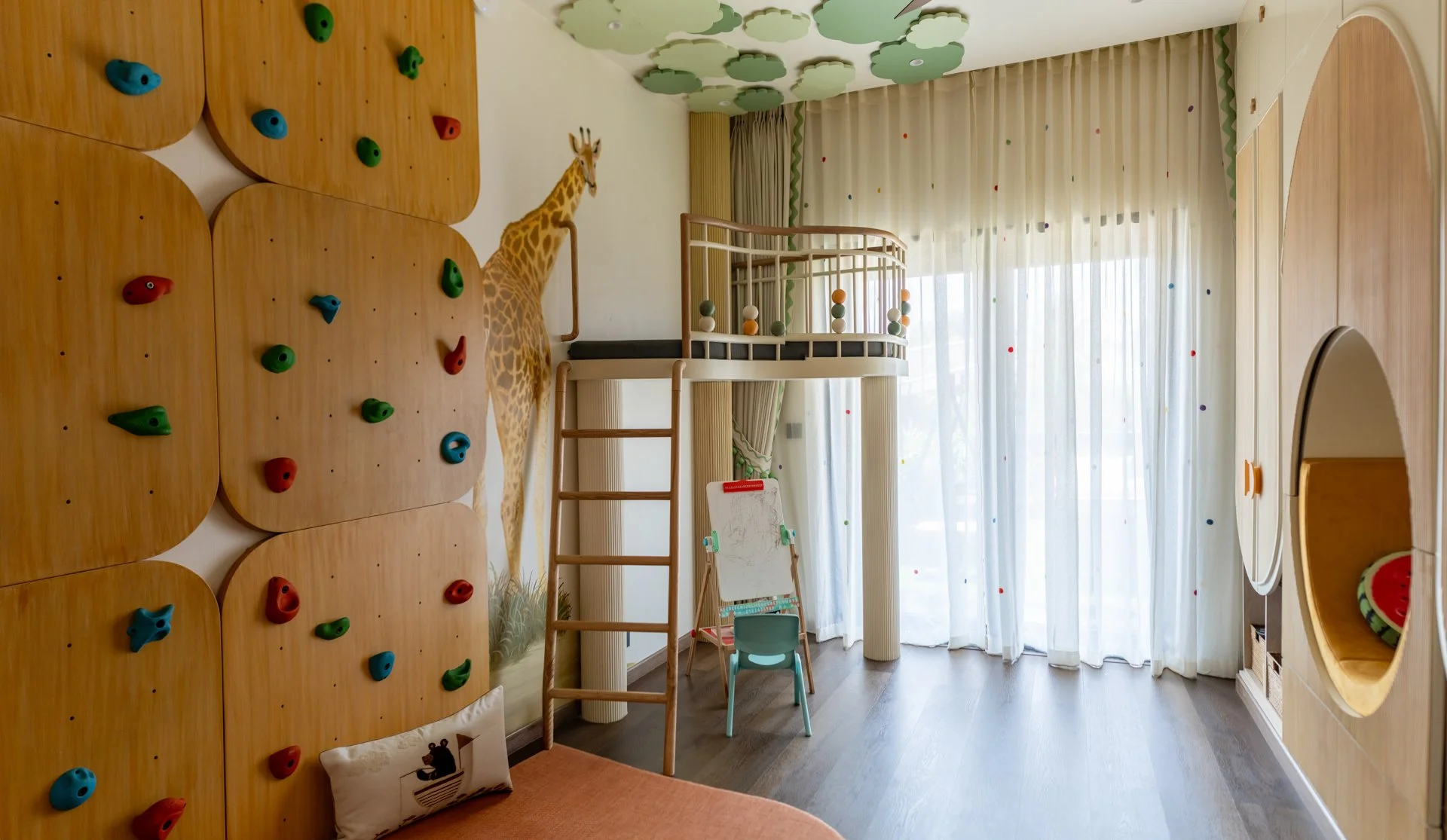 Colorful indoor playroom with climbing wall, a giraffe figure, a loft play area with a safety rail, a children's easel, and a window with sheer curtains decorated with colorful dots.