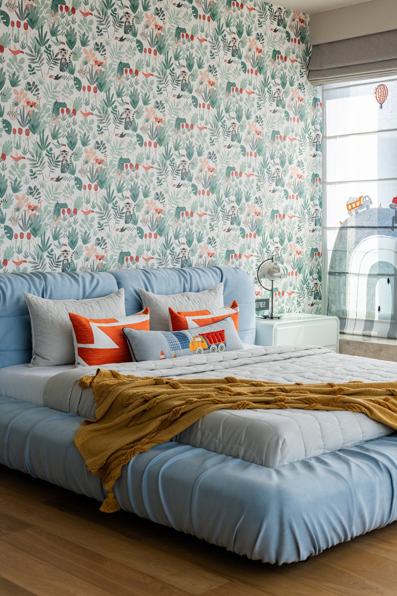 A bedroom with a light blue upholstered headboard, white pillows with orange accents, a brown blanket, and a wallpaper featuring jungle animals and plants. There is a window with a roller shade and a wall mural of a road with construction vehicles.