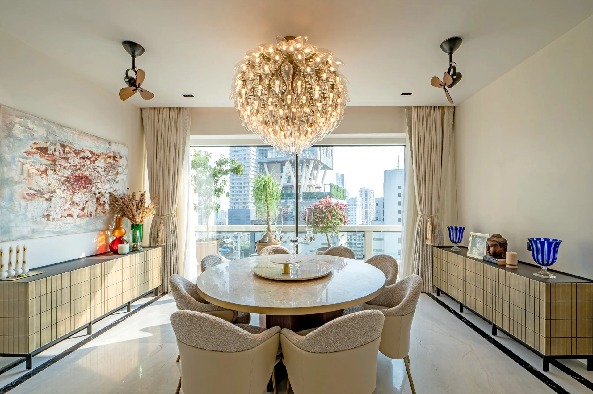 Modern dining room with a marble table surrounded by beige upholstered chairs, a chandelier overhead, and a large window showing city buildings outside. Decor includes vases, candles, and artwork.