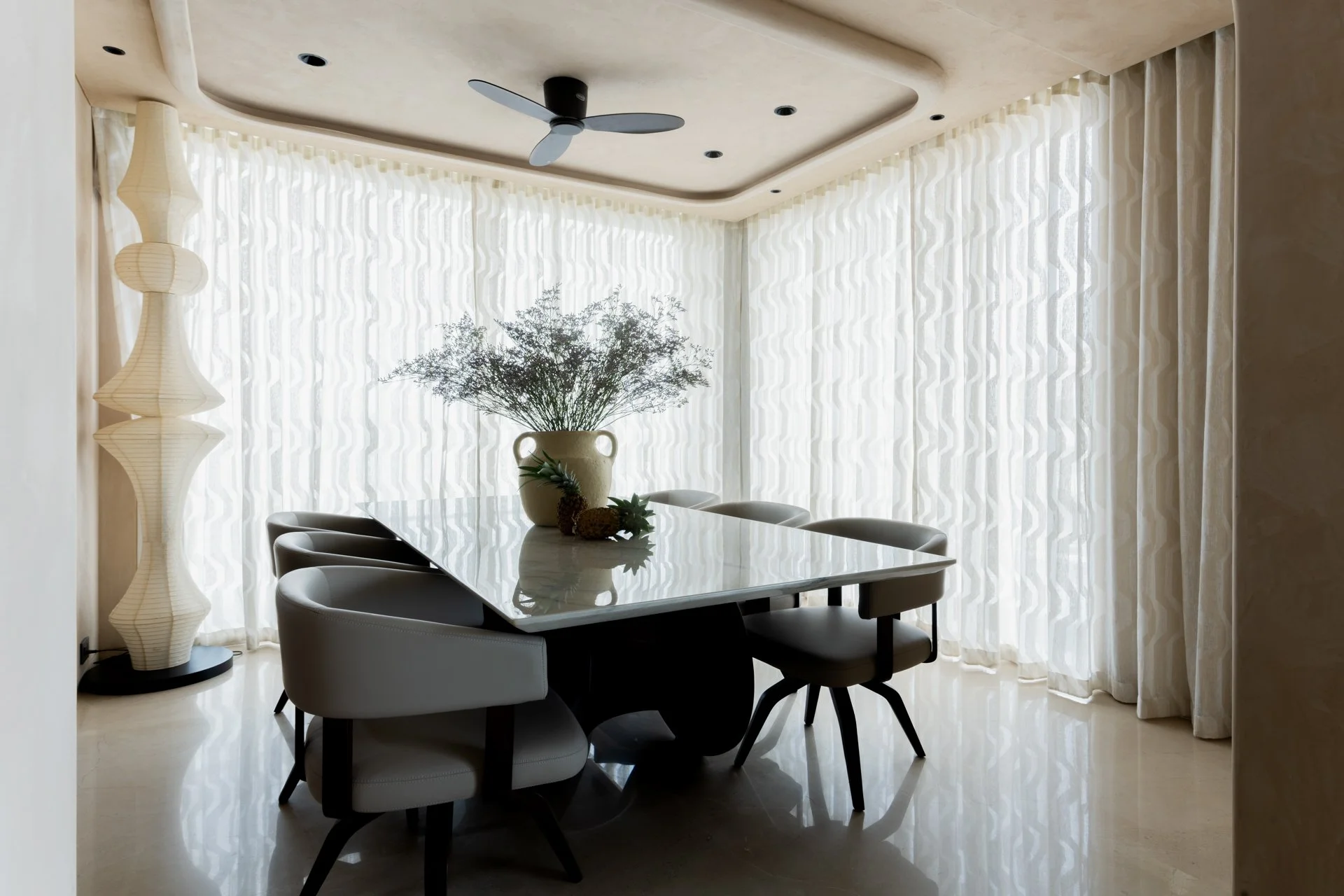 Dining room with a white marble table, six chairs, a large vase with greenery, a decorative floor lamp, sheer curtains, and a ceiling fan.