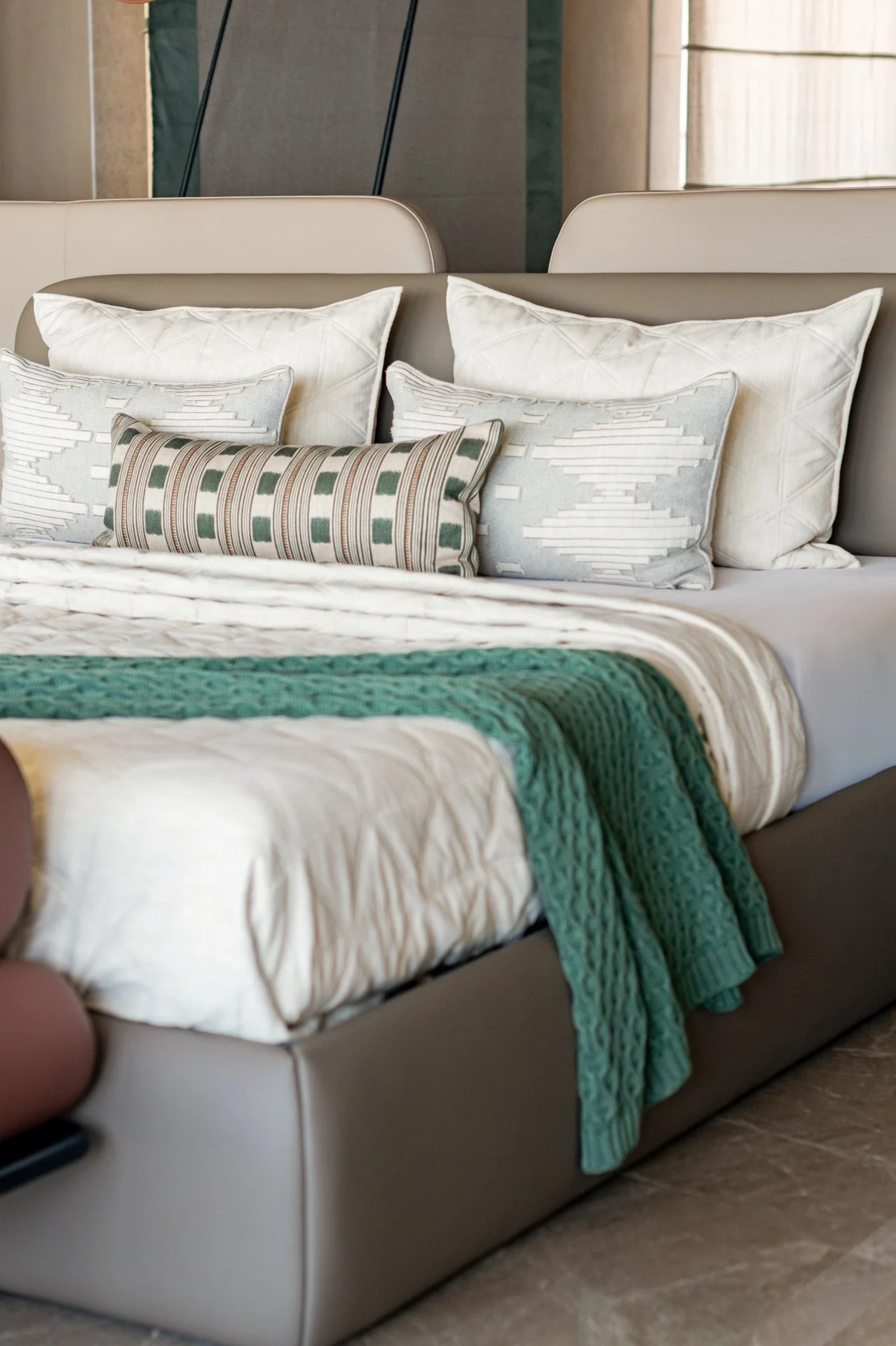 A bed with multiple pillows, including white pillows with diamond stitching, decorative pillows with geometric patterns, and a textured green blanket at the foot. The headboard is upholstered in a neutral color, and the setting appears to be a hotel or modern bedroom.