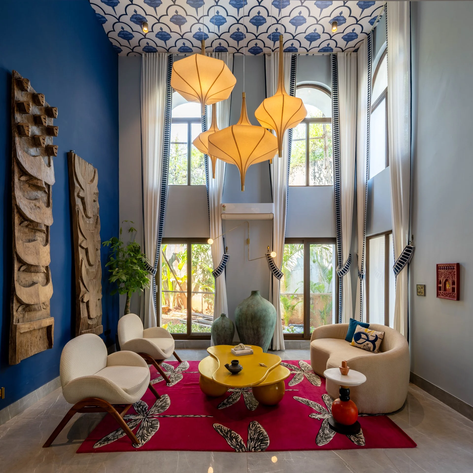 A bright living room with large windows, white curtains with black trim, and a high ceiling with patterned blue and white wallpaper. The room features modern furniture, including white chairs, a beige curved sofa with blue and white pillows, a yellow coffee table, and decorative vases. A red rug with black and white floral patterns is on the floor. There are hanging pendant lights with beige shades and wooden accents.