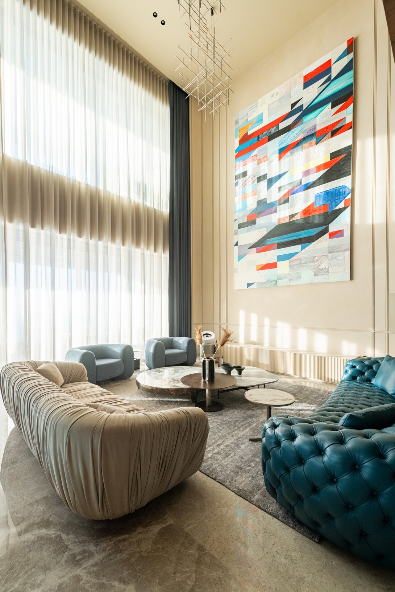 Modern hotel lobby with large abstract painting on the wall, high ceiling, floor-to-ceiling curtains, and eclectic seating including a beige striped sofa, blue tufted armchair, and small round tables.