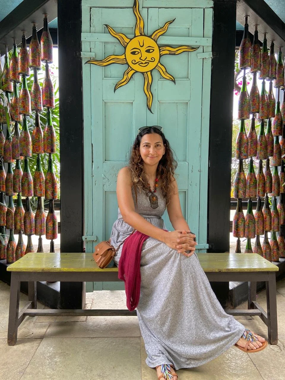 A woman sitting on a bench indoors, wearing a long silver dress with a red scarf, smiling, with a decorative sun with a face on the wall behind her, and colorful curtains with bottles pattern on each side.