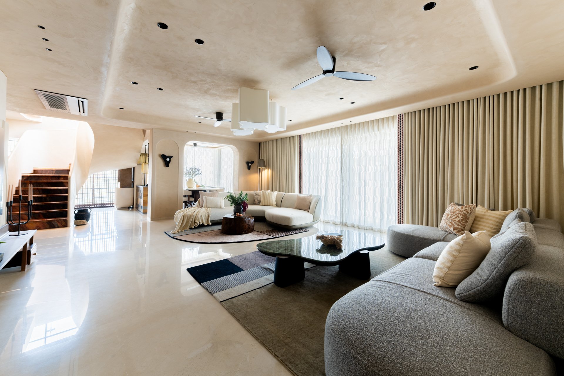 Living room with beige walls, cream-colored curtains, and modern furniture including sofas, a coffee table, and a rug. There are ceiling fans and recessed lighting, with a staircase visible in the background.