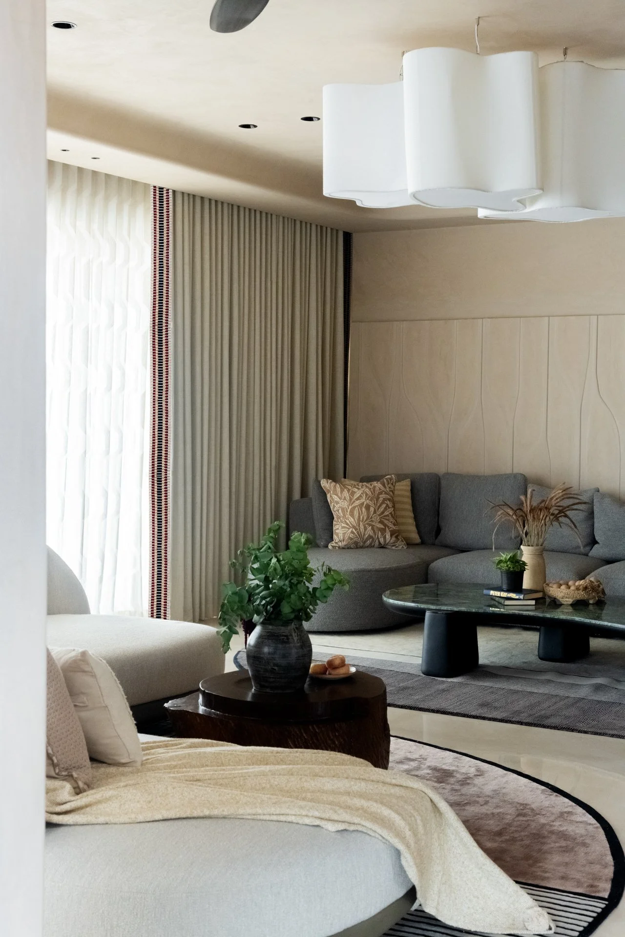 A modern living room with a gray sectional sofa decorated with patterned throw pillows. There is a round black coffee table with a plant, books, and a decorative bowl. A white ceiling light fixture hangs above. Light-colored curtains cover the window, and a beige wall with a subtle pattern is on the right side.