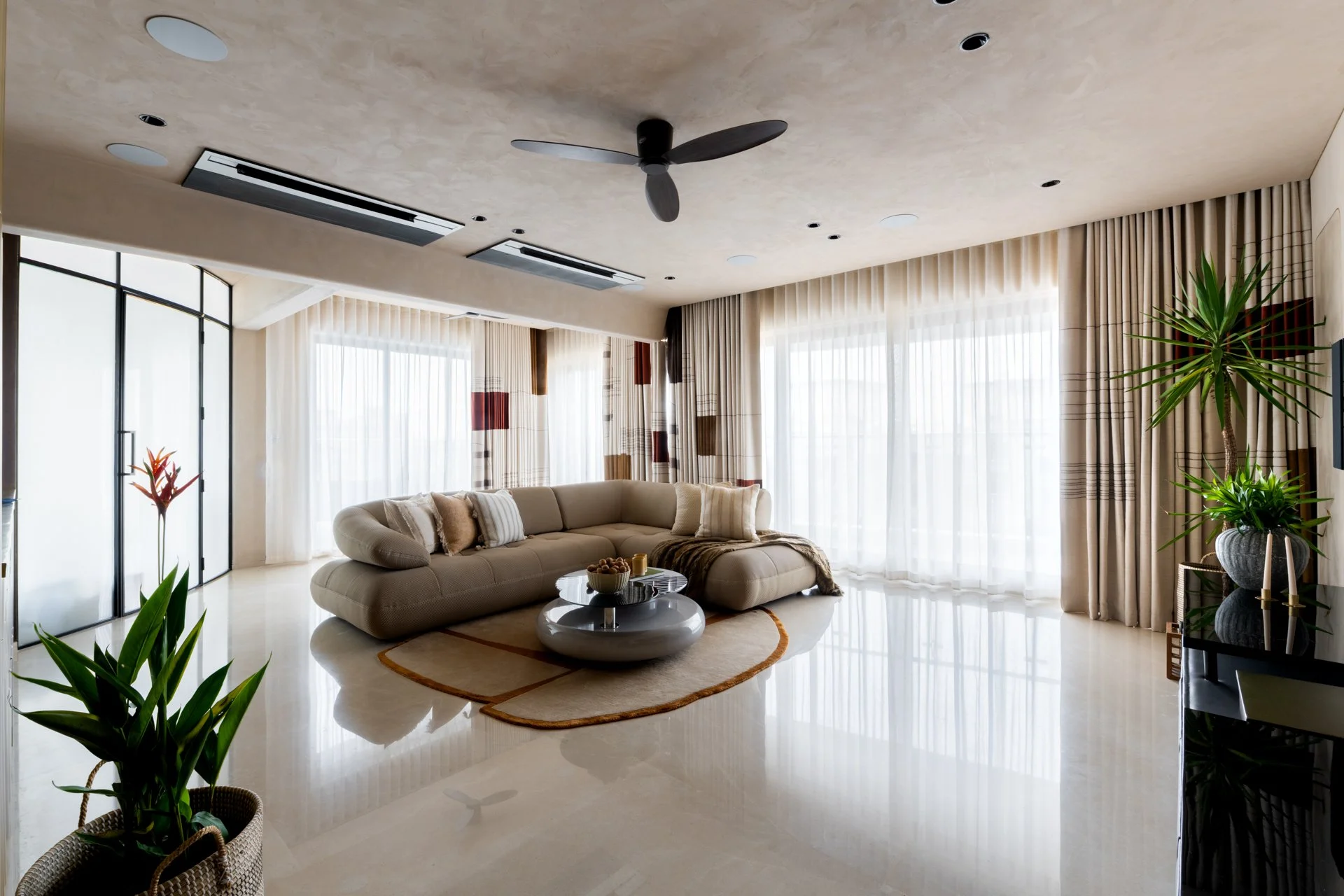 Modern living room with white curved sofa, round coffee table with bowls, large windows with sheer curtains, indoor plants, ceiling fan, and recessed lighting.