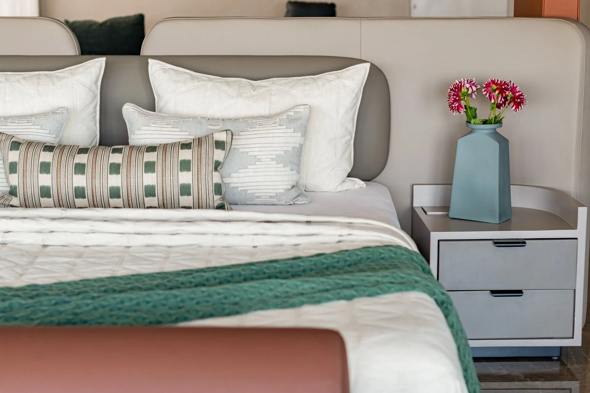 Close-up of a bed with multiple pillows, including white and patterned decorative pillows, and a bedside table with a blue vase holding pink and white flowers.