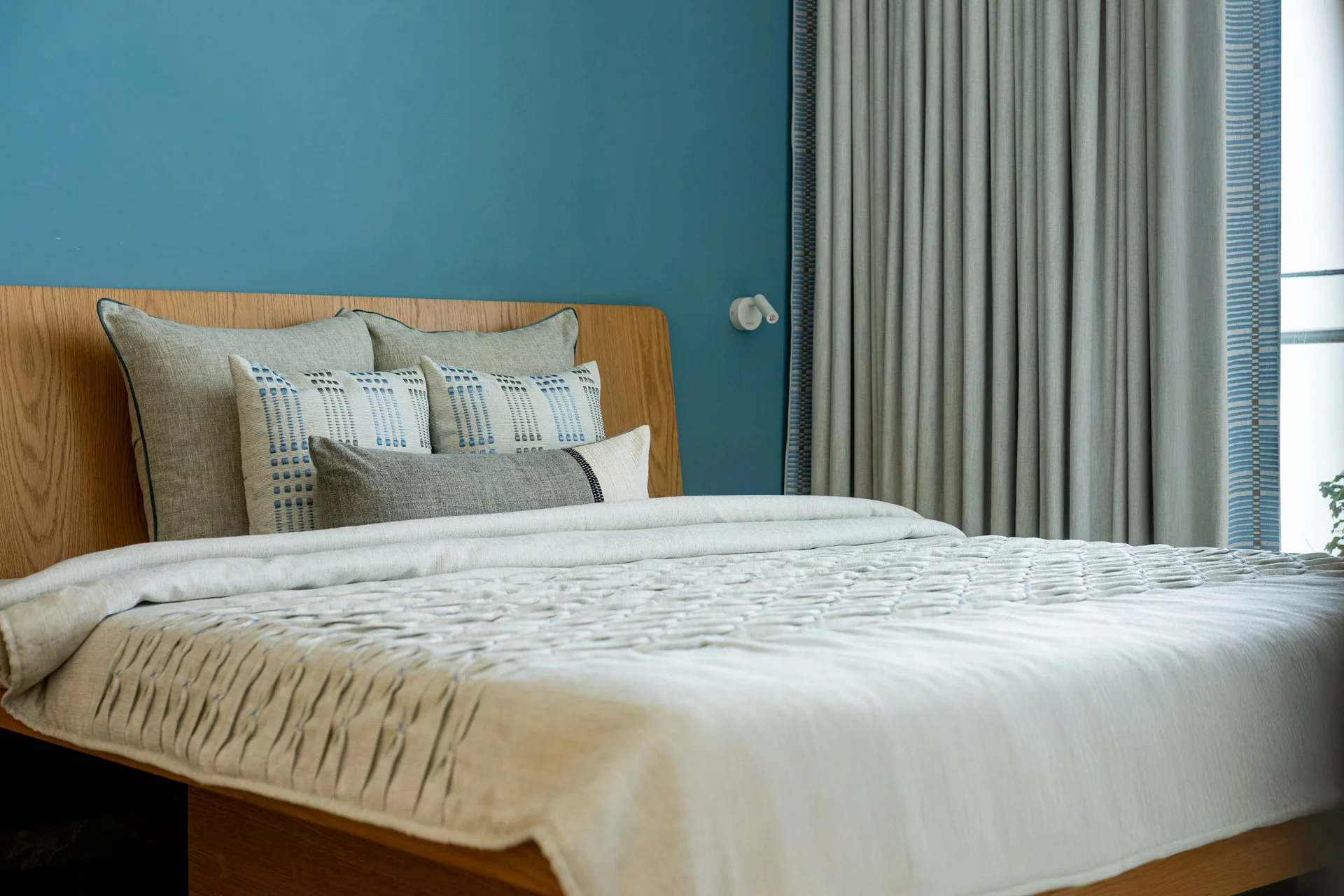 A neatly made bed with multiple pillows against a wooden headboard, set in a room with a blue wall and closed curtains on a window.