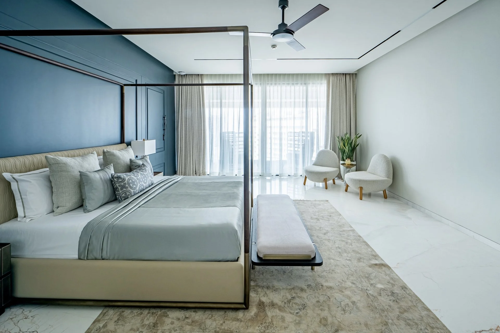 Modern hotel room with a bed, light-colored bedding and pillows, a small chair, a side table with a plant, large window with curtains, and a ceiling fan.